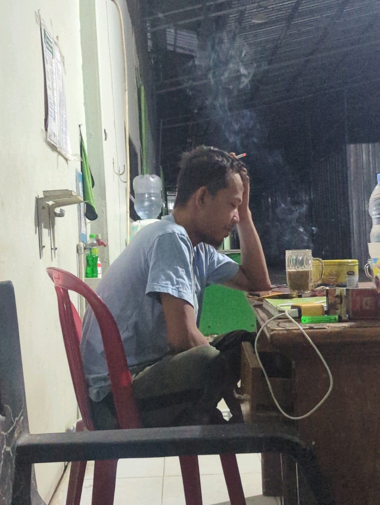 A man is seated at a table, smoking a cigarette, with his head resting on his hand. He is wearing a light blue t-shirt and dark pants. In front of him on the table is a glass of coffee, a yellow tin, and what appears to be a book or notebook. A green lighter is also visible on the table. A water dispenser is in the background, along with some shelves and a bottle of green liquid. The scene appears to be indoors, possibly in a home or small business in Batu Belaman, Indonesia, given the context.