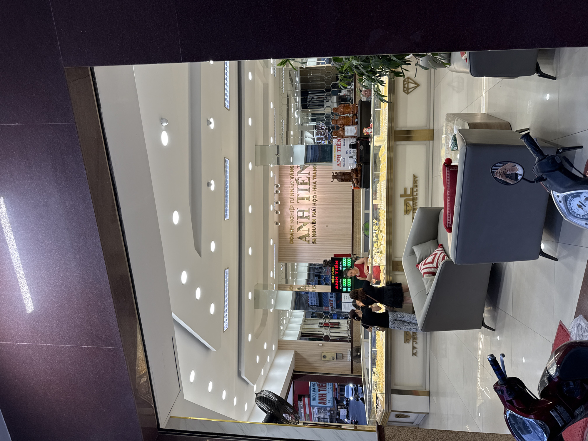 The image depicts the interior of Anh Tien Jewellery store, located at 58 Nguyen Thai Hoc Street in Nha Trang, Vietnam. The store features a well-lit interior with a display of gold jewellery behind a glass counter. Several individuals are present, including two women standing at the counter examining the jewellery, and a woman in a red dress, likely an employee, assisting them. Another older woman is partially visible behind the counter. In the foreground, a motorcycle is parked, and a seating area with grey sofas and a coffee table is visible. The store's signage, including "Doanh Nghiep Tu Nhan Vang Anh Tien" and "58 Nguyen Thai Hoc - Nha Trang," is prominently displayed. Outside the store, a street scene with a fan and vehicles can be observed through the entrance.
