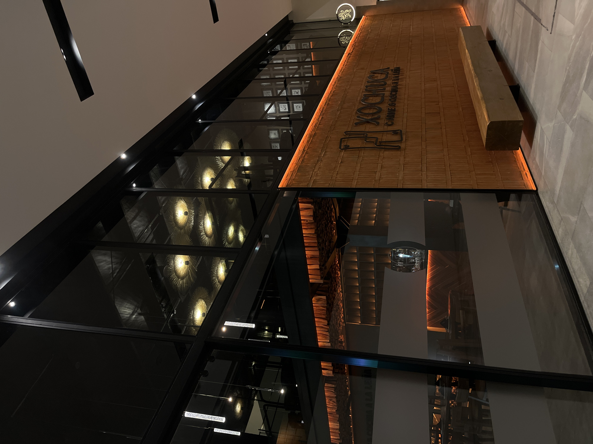 The image shows a building with a modern architectural design, featuring a glass facade framed by black metal beams. Inside, circular, decorative lights can be seen, suggesting an interior space. Above the glass, there is a wooden wall with the name "XOCHOIICA" and the phrase "CARNE & COCINA LA LEÑA" inscribed on it, indicating a restaurant or similar establishment. A solid rectangular block is mounted on the wall next to the writing. The surrounding environment appears to be a commercial area, possibly in the city of San Bernardino Tlaxcalancingo, Mexico.