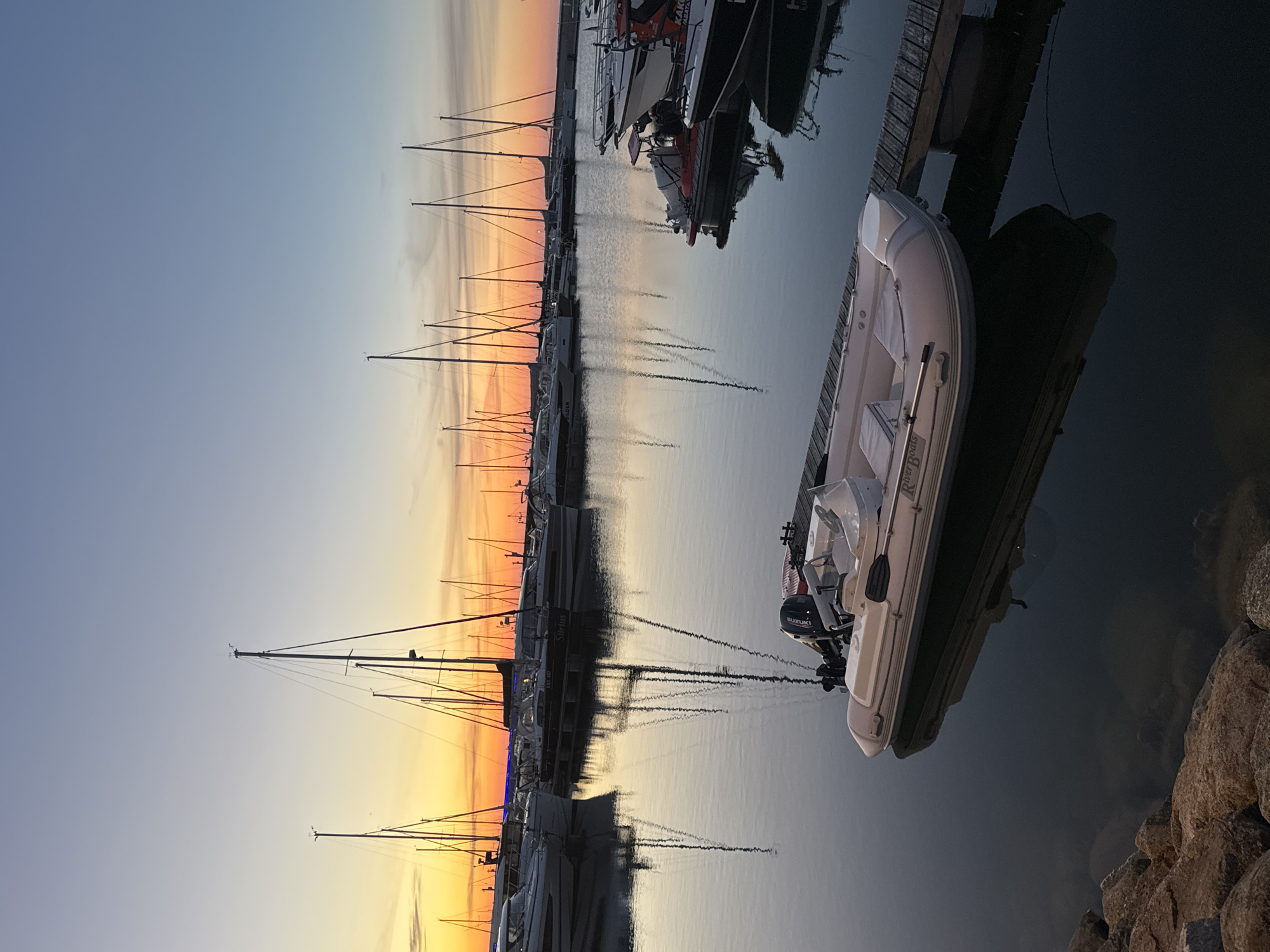 A still shot of a marina in Tallinn, Estonia at sunset. The sky is a gradient of orange, yellow, and blue. Several sailboats are docked in the background, their masts creating a silhouette against the colorful sky. In the foreground, a white inflatable boat with a small outboard motor is tied to a wooden dock. The water is calm and reflects the boats and the sky. Rocks are visible in the lower left corner of the frame. There are no people in the image.