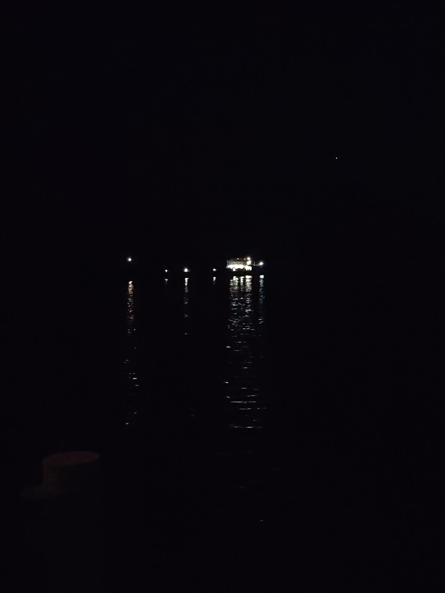 The image shows a dark scene with light reflecting off the water. In the distance, there appear to be lights from a structure or ship, possibly in a body of water like a river or lake. Closer to the foreground on the left, there is a dark shape. A single bright spot appears in the sky. The location is Unknown.