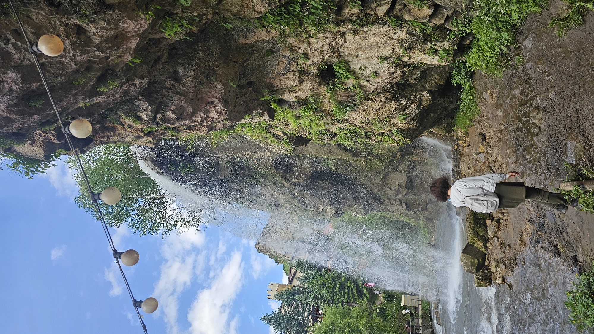 In the city of Krasny Kurgan, Russia, a young person with dark, curly hair and a light gray backpack is standing on a muddy path, facing a waterfall. They are wearing a light gray sweatshirt and an olive-green skirt or dress over olive-green leggings or tights. The waterfall cascades down a large, rocky cliff face that is covered in green vegetation. In the background, to the left of the waterfall, are evergreen trees and a building with a tower, resembling a castle. Above the scene, a string of round, warm-toned lights is strung across the frame against a bright blue sky with scattered white clouds. The ground in front of the waterfall is a mix of rocks and mud, with a stream flowing to the left.
