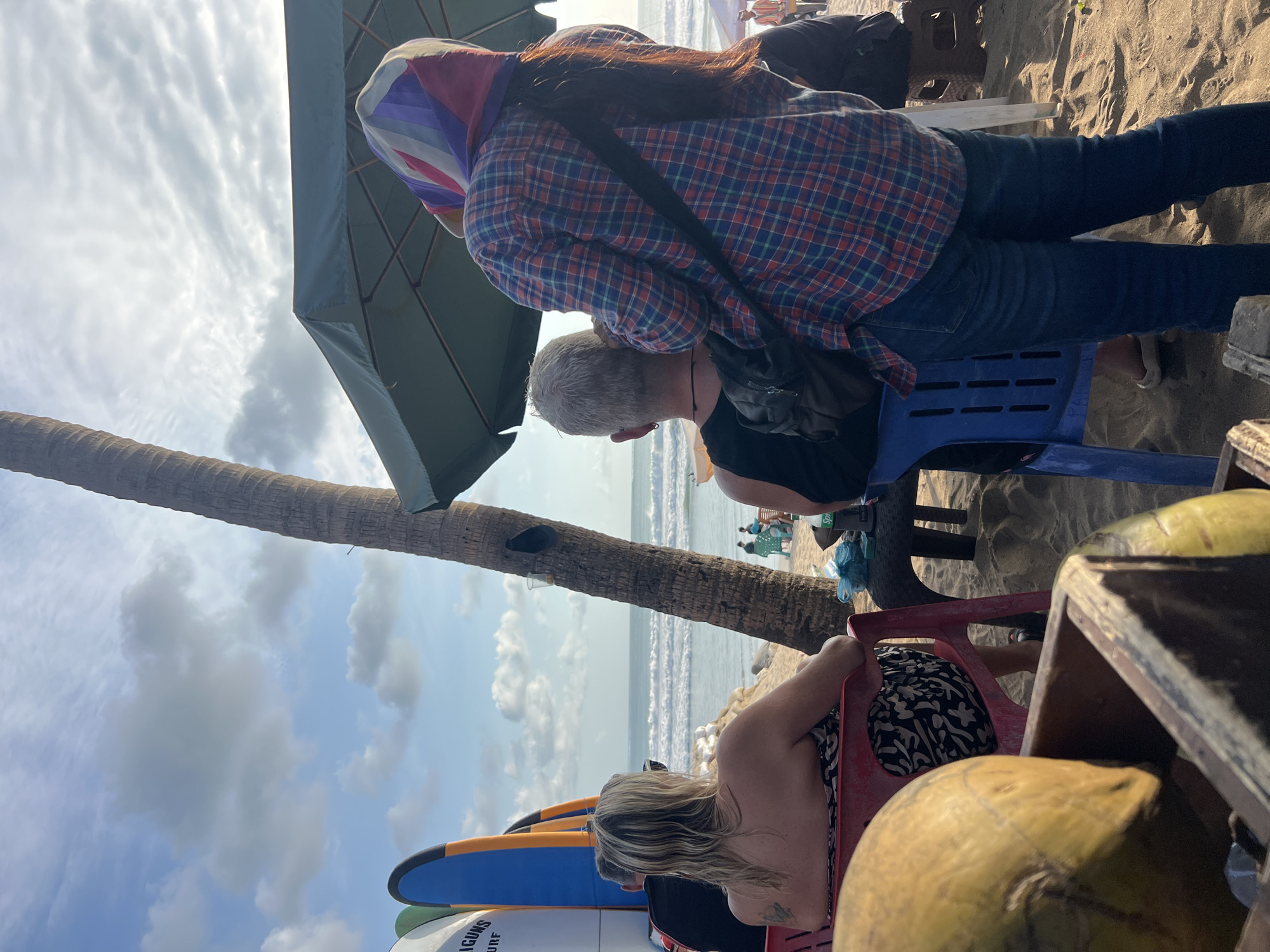 In Kuta, Indonesia, a beach scene unfolds under a partially cloudy sky. In the foreground, a wooden crate holds two coconuts, one prominently in focus. Behind the coconuts, a woman with blonde hair sits in a red plastic chair, facing away from the camera, looking towards the ocean. She wears a patterned strapless dress. Beside her, a man with short grey hair sits in a blue plastic chair, also facing the ocean, wearing a black tank top. A woman in a red and blue plaid shirt and blue jeans stands behind him, facing away, wearing a colorful headscarf and a black crossbody bag. A palm tree trunk stands vertically in the mid-ground. Beyond the people, surfboards are propped up on the sand to the left, with the ocean and waves visible in the distance. Beach umbrellas and other beachgoers are scattered further down the shoreline. The sand is a light brown, and the water appears turquoise with white crests on the waves. The overall atmosphere is relaxed and suggests a day at the beach.