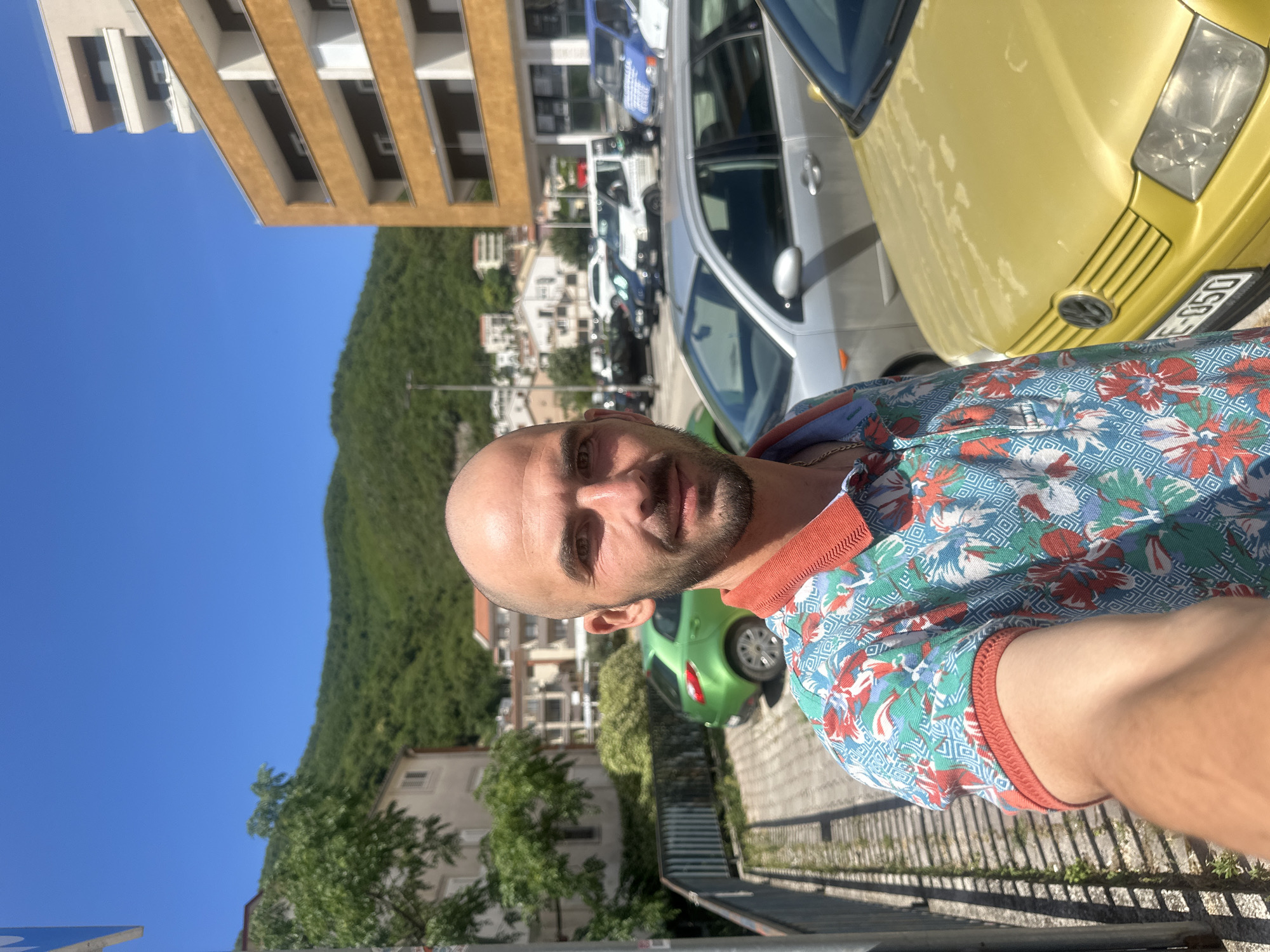A bald man with a short beard is captured in a bright outdoor selfie, looking directly at the camera with a neutral to slight smile. He wears a vibrant short-sleeved shirt featuring a tropical floral and geometric pattern in shades of blue, red, and white, with an orange-red collar and sleeve trim.
The background reveals a sunny day with a clear, vivid blue sky dominating the upper left portion of the frame. A lush, steep, green hill or mountain, densely covered in trees, rises in the middle ground. Various buildings are visible: a multi-story modern structure with a brown facade and white balconies stands to the far left, while smaller white and tan buildings with terracotta roofs are scattered across the hillside.
Several cars are parked along what appears to be a street or parking area. A prominent yellow car, likely a Volkswagen, is partially visible to the man's right, showing its front bumper and headlight. A fragment of its license plate, "E-050", is discernible. Further back, a silver sedan and other vehicles, including a white van and a blue car, are parked. Another bright green car is visible behind the man's head. The scene suggests a warm, clear day in an urban or semi-urban environment, consistent with the provided location context of Budva, Montenegro.