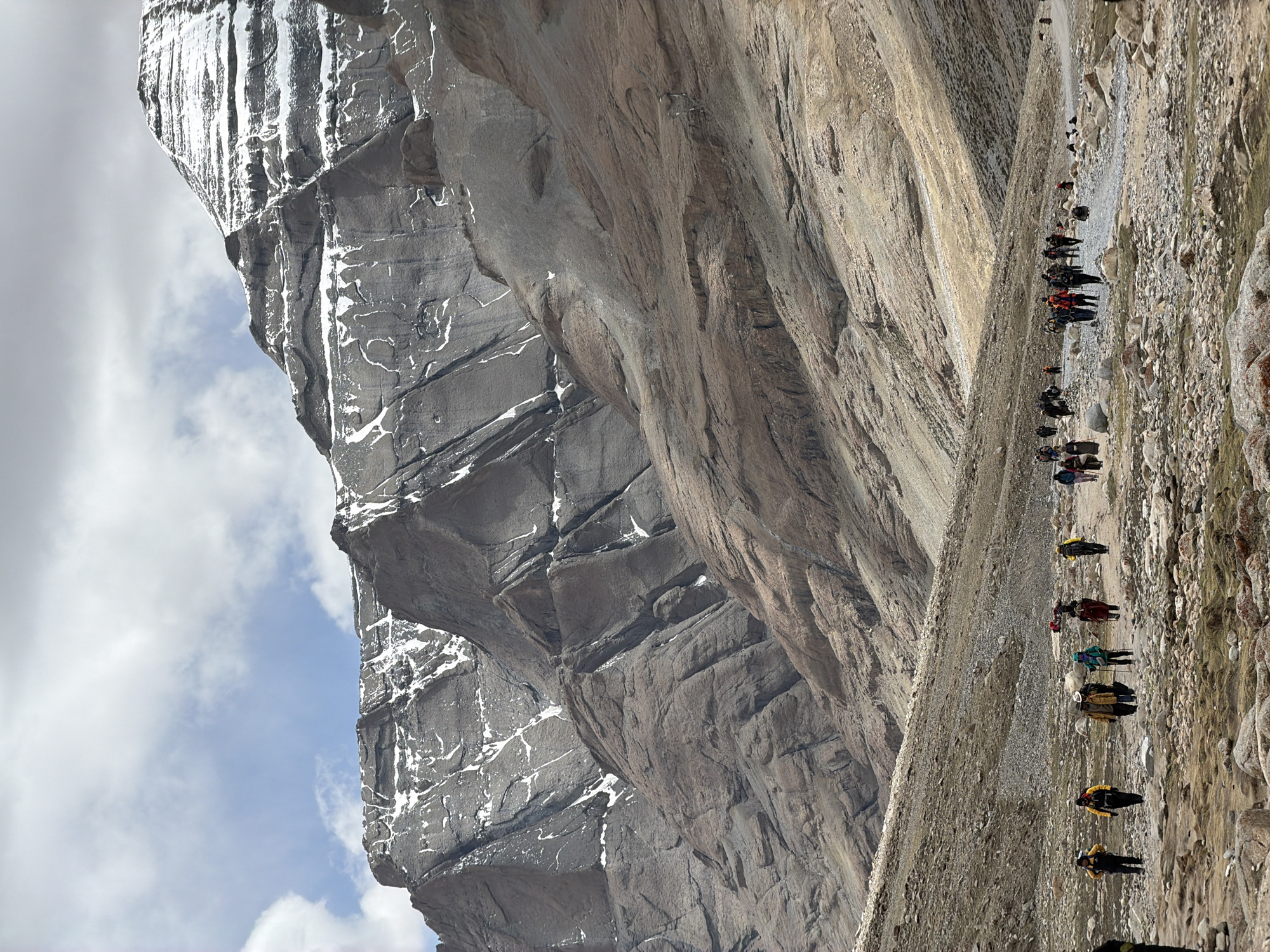 The image shows a mountain scene in what appears to be a remote area. A group of people and pack animals is moving along a rocky path at the base of a towering cliff. The mountain is composed of different colored rock layers, with some sections showing signs of snow or ice. The sky is partially cloudy. There is no evidence to support the location being in Parga town, China.