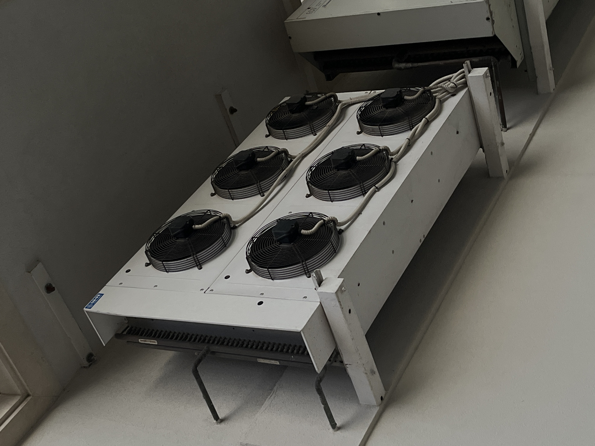 This is a low-angle, eye-level shot of industrial cooling equipment mounted on a light-colored wall, likely in Budva, Montenegro. The main subject is a large, white, rectangular unit with six black industrial fans arranged in two rows of three. The fans are protected by black grilles, and white protective tubing snakes around them, connecting to the unit. The unit is supported by white metal brackets attached to the wall. To the right of the main unit, a portion of another white industrial unit is visible, with black vertical elements suggesting internal components. The background consists of a dark, possibly textured ceiling and a light-colored wall. The overall impression is one of functional, outdoor industrial equipment.
