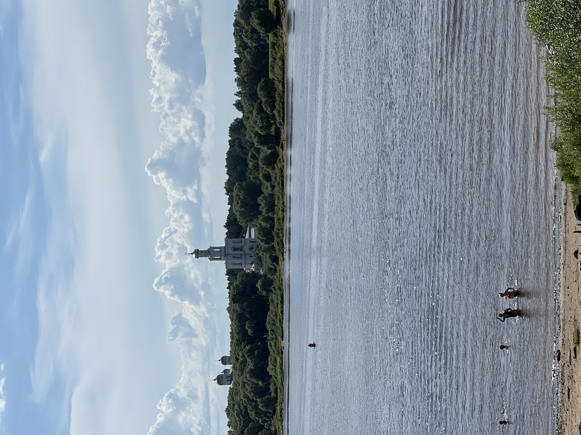 The image captures a wide, calm river in Veliky Novgorod, Russia, with a sandy bank in the foreground. Several individuals are visible in the water, some appearing to be swimming or wading. In the mid-ground, a red buoy marks a spot in the river. Across the water, a dense line of green trees forms the horizon, behind which rise several white church buildings with distinctive golden domes. A tall, slender bell tower is particularly prominent. The sky above is a vibrant blue, dotted with fluffy white clouds. The overall scene conveys a sense of a warm, sunny day with recreational activity on the river.