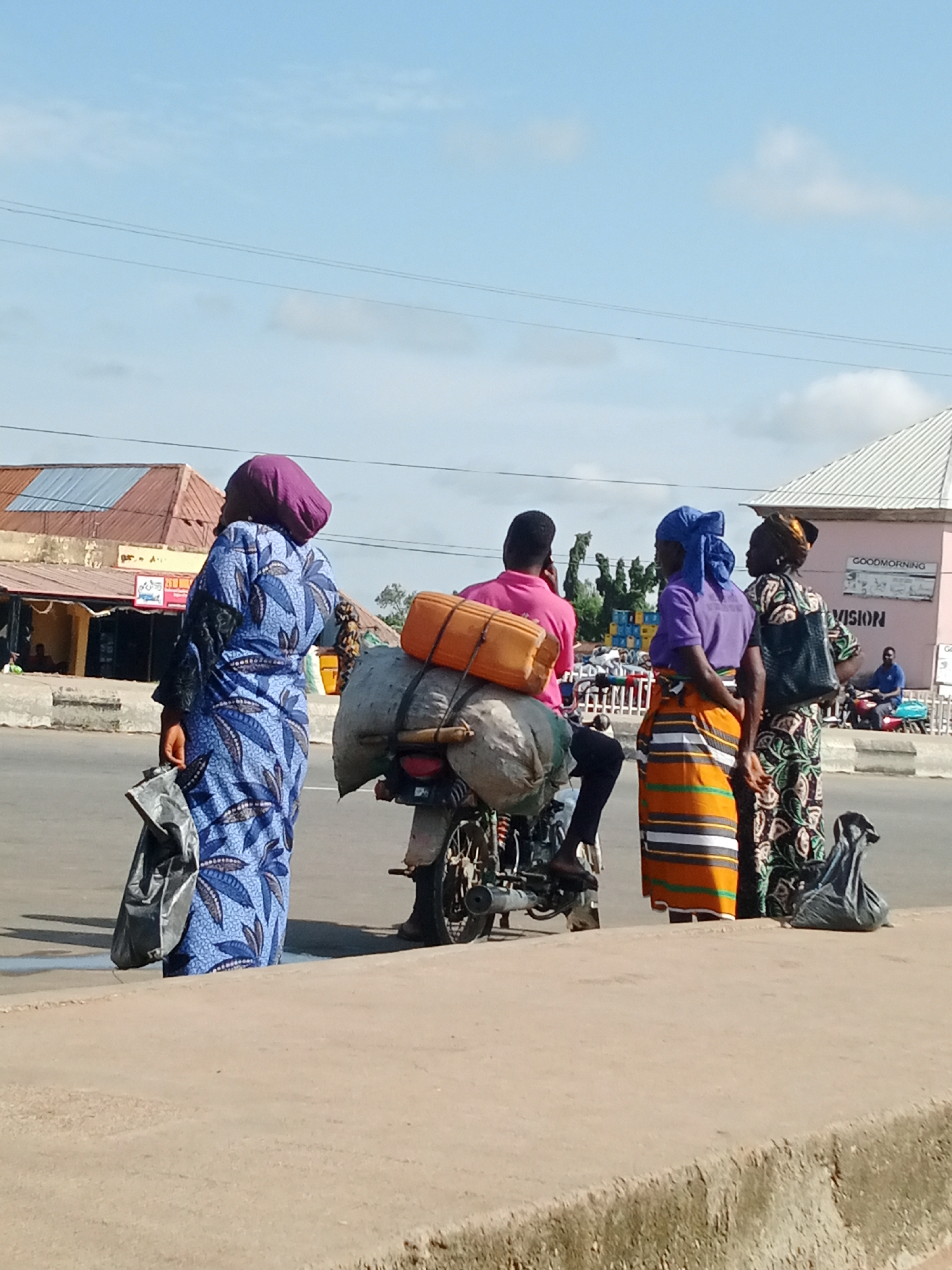 In a street scene in Unknown, Unknown, four individuals are gathered around a motorcycle that is loaded with two large sacks. On the far left, a person wearing a blue and white patterned dress and a purple headscarf stands holding a black plastic bag. To their right, a person in a pink shirt sits on the motorcycle, facing away from the camera. A person in a purple shirt and a colorful striped skirt stands to the right of the motorcycle driver, facing right. Behind them, a person in a black, green, and white patterned dress and a headscarf is partially visible, also facing right. The motorcycle is parked on a paved road, with buildings visible in the background. One building has a sign that reads "GOODMORNING VISION". The sky is clear with a few scattered clouds.