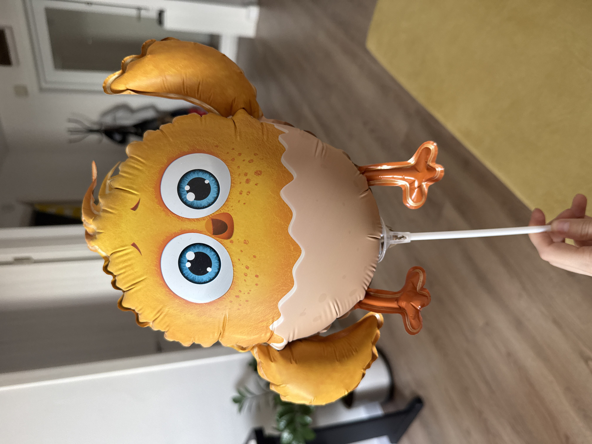 An eye-level, medium shot depicts a yellow and orange chick-shaped balloon with large blue eyes, held by a person. The background reveals an interior space with white walls, a door frame, a black object that could be a plant stand or other furniture, and a light-colored wooden floor. Part of a yellow sofa can be seen on the right side of the image. There is insufficient information in the provided image to determine whether the location is in the city of Khimki, Russia.