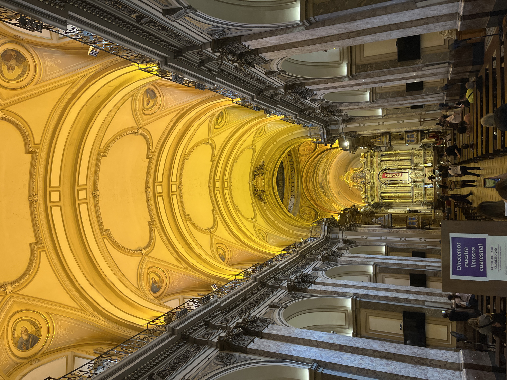 The image captures an interior view of a cathedral in Buenos Aires, Argentina. The camera is positioned looking upward, emphasizing the architectural details of the ceiling and the columns lining the space. The ceiling features a series of arches with intricate detailing and portraits within circular frames. The predominant color is a warm yellow, likely from interior lighting. Along the sides, marble columns support the structure, with ornate detailing at the top. A wrought iron balcony runs along the side, adding another layer of architectural interest. People are seated in pews, presumably for a religious service. In the foreground, there is a sign in Spanish that reads "Ofrecemos nuestra limosna cuaresmal" which translates to "We offer our Lenten alms," referring to the Lenten season in Christianity. The sign also mentions "GESTO SOLIDARIO CUARESMA 2026" indicating a Lenten campaign or initiative related to solidarity. The overall scene conveys a sense of grandeur and reverence within the religious setting.
