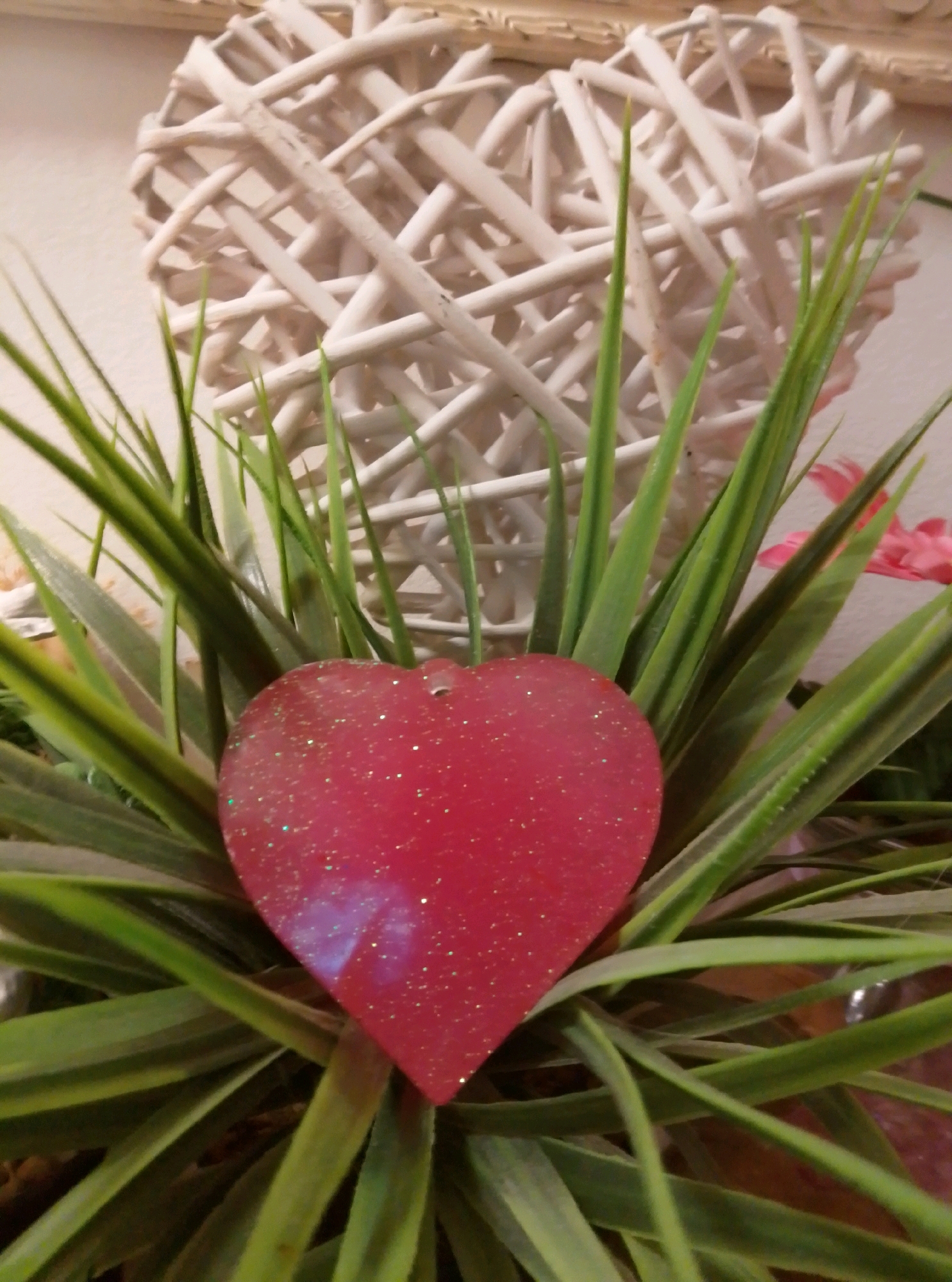 A close-up, head-on shot shows a bright pink, heart-shaped glitter ornament resting on a bed of green, spiky artificial grass. Behind the heart, a large, woven white heart-shaped decoration is partially visible against a light-colored wall. The scene is indoors, and the overall lighting is warm and soft. The image does not provide any information about the city of Wedel, Germany, as it depicts a close-up of decorative objects.