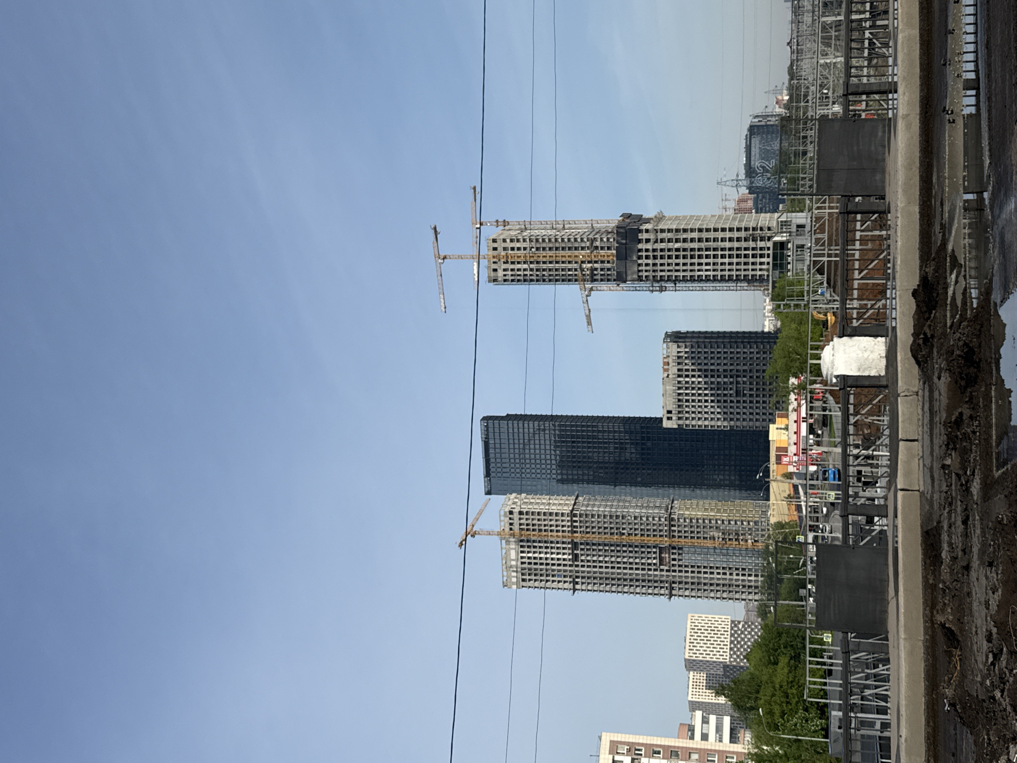 The image depicts a cityscape in Moscow, Russia, with a focus on construction sites. Several high-rise buildings are under construction, characterized by their skeletal structures and the presence of cranes. In the foreground, there is a concrete structure, likely a wall or barrier. Power lines stretch across the sky. The environment appears to be urban, with a mix of modern and older buildings visible in the distance. The sky is clear and blue. No people are visible in the immediate view.