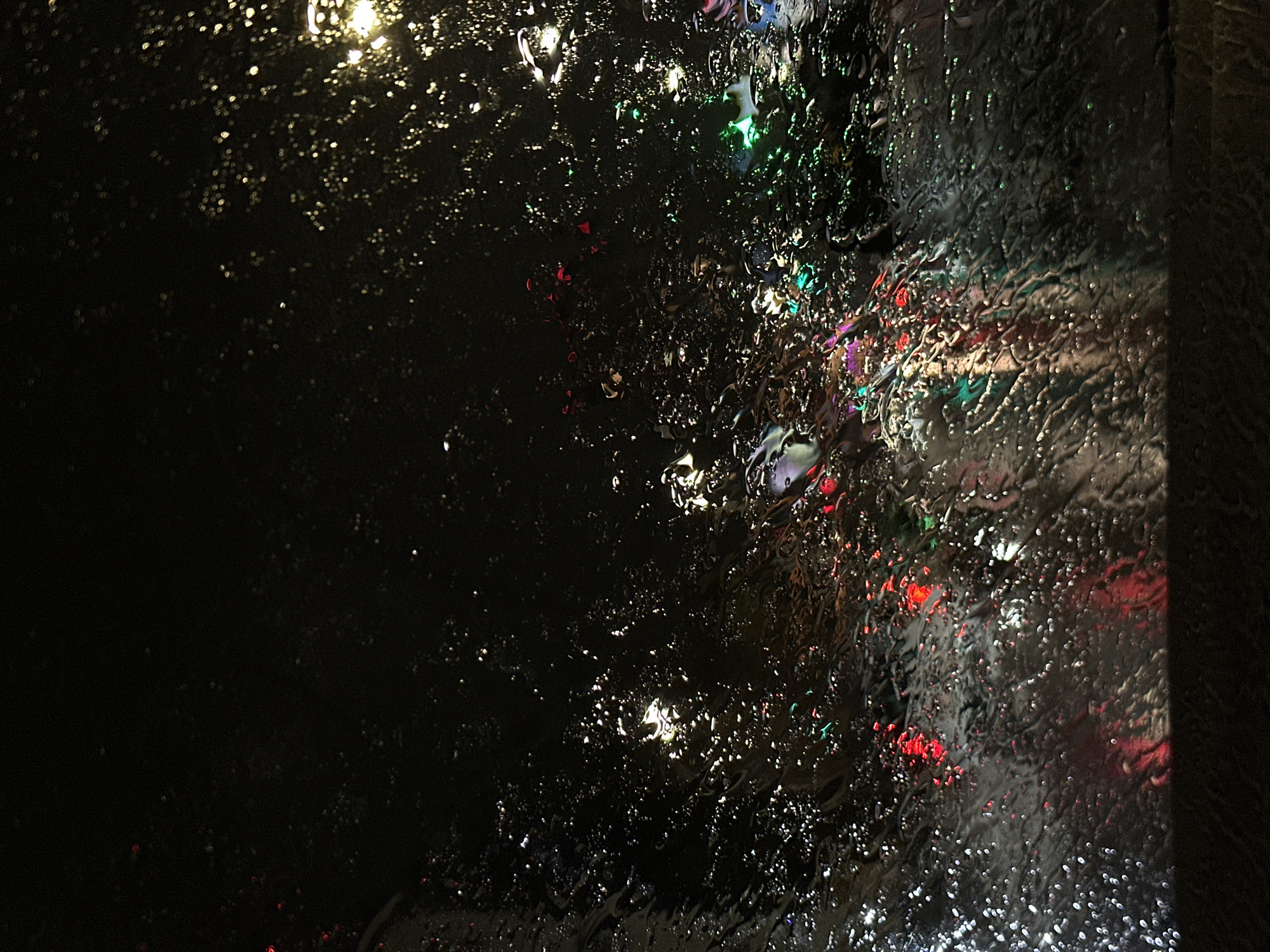 A close-up view through a rain-streaked window at night. Streaks of water obscure lights of various colors, including white, red, and green. The right edge of the window frame is visible. Most of the window is dark. The location is likely a city street based on the colored lights.