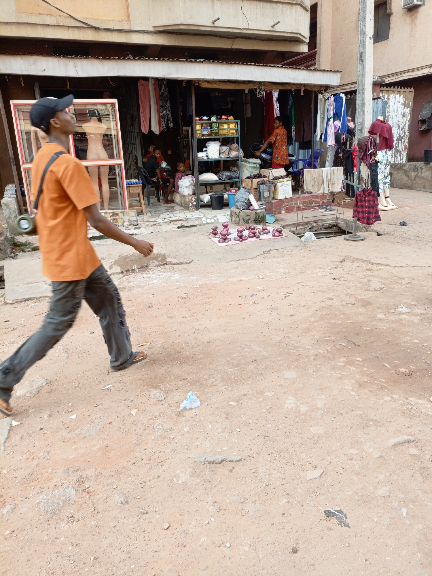 The image depicts an outdoor market or shop front during the daytime, characterized by a bustling and informal atmosphere. In the foreground, a person walks from left to right, their body slightly blurred, indicating movement. They wear an orange short-sleeved shirt, dark grey or black distressed pants with visible wear, brown sandals, and a black baseball cap. A cylindrical object, possibly a portable speaker or large flask, is slung over their shoulder with a black strap. The ground is unpaved, consisting of reddish-brown dirt and small stones, with scattered litter including small pieces of plastic.
Behind the walking figure, an open-fronted shop or stall is situated beneath a corrugated metal awning. To the left, a large, empty window frame with a red trim stands upright, partially obscuring a female mannequin torso inside the shop. The shop interior features shelves stocked with various packaged goods. Several individuals, including children, are visible sitting within the shop area. Clothes are displayed both inside the shop and hanging from racks and lines outside. Notably, a red and black plaid shirt hangs on a freestanding display to the right of the main shop entrance. Further right, a mannequin displaying pants and a patterned top stands on white boots.
On the ground directly in front of the shop, a pile of reddish-purple items, resembling onions or similar root vegetables, is neatly arranged on a mat. Nearby, a white rectangular container with the word "LUCI" printed on it is visible. Various buckets and other containers are also scattered around the shop's perimeter. The background shows parts of multi-story buildings, suggesting an urban or semi-urban environment.