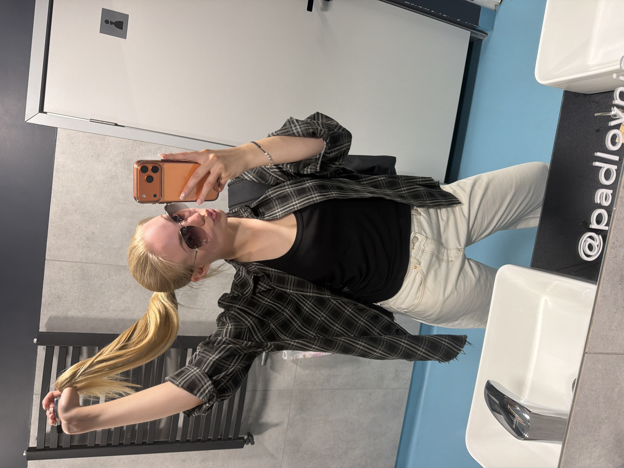 A young, light-skinned person with blonde hair in a low ponytail is taking a selfie in a public restroom. They are wearing sunglasses, a black tank top, a grey and black plaid shirt, and white jeans. The person is holding a copper-colored smartphone with their right hand. The restroom has grey tile walls, a white door with a gender sign, a black radiator, a white sink, and a blue accent wall. The text "@padlovyn" is visible on the mirror near the sink. The location is Unknown, Unknown.