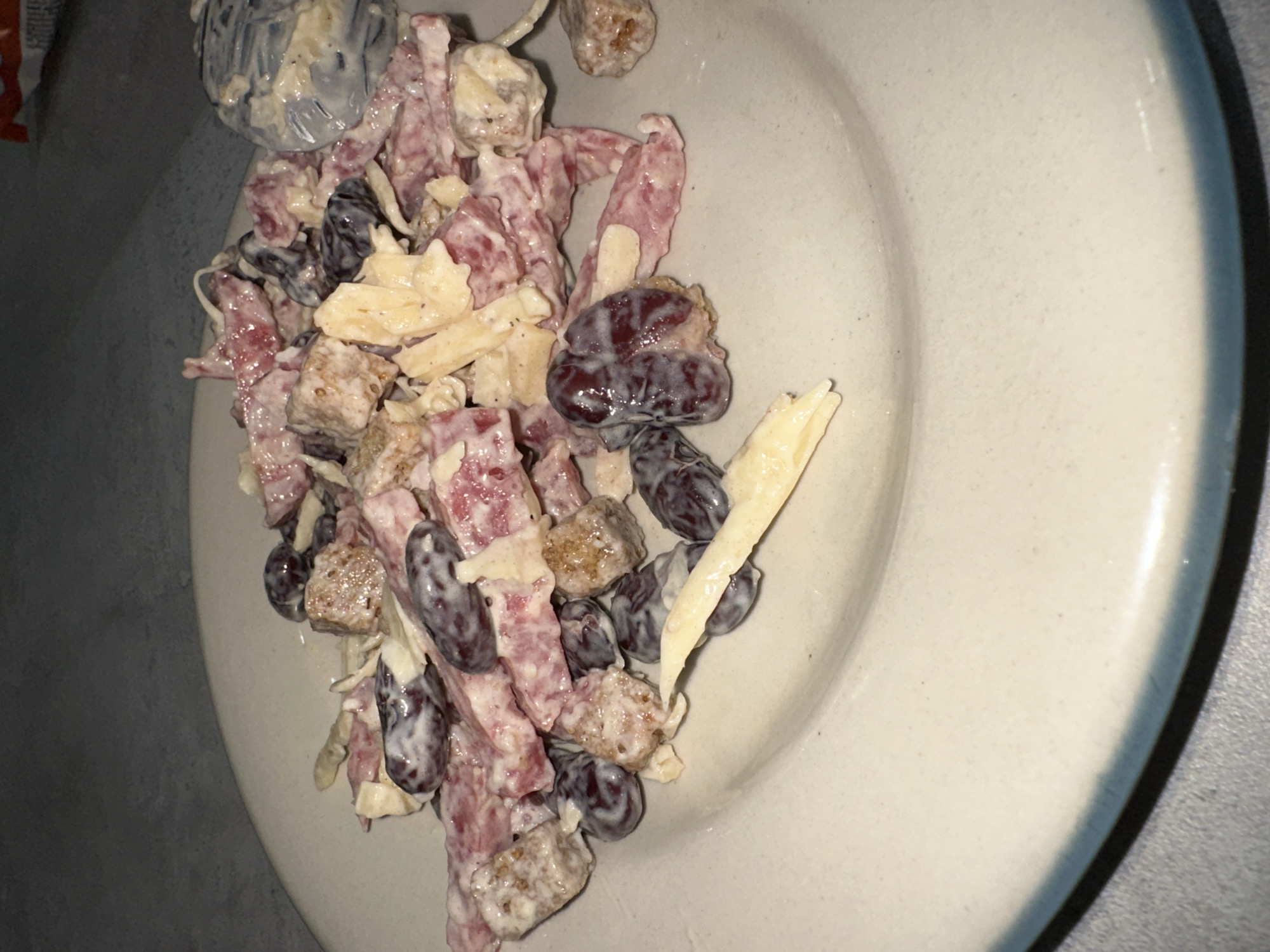 The image displays a close-up, overhead view of a creamy dish served on a light-colored, possibly ceramic plate with a visible rim. The dish features a mix of ingredients heavily coated in a white, mayonnaise-like dressing. Key components include reddish-pink strips, dark red to purple oval shapes resembling grapes or kidney beans, irregular yellowish pieces, and small, light brown cube-like chunks, likely croutons or bread. A spoon with a blue-grey handle, also covered in the dressing, is partially visible in the upper left corner, suggesting the dish is ready for consumption. The background is a dark, undifferentiated surface, likely a tabletop, with a small, blurry orange object visible on the far left. The scene depicts an indoor meal. The image was captured in Chaikovsky, Russia.