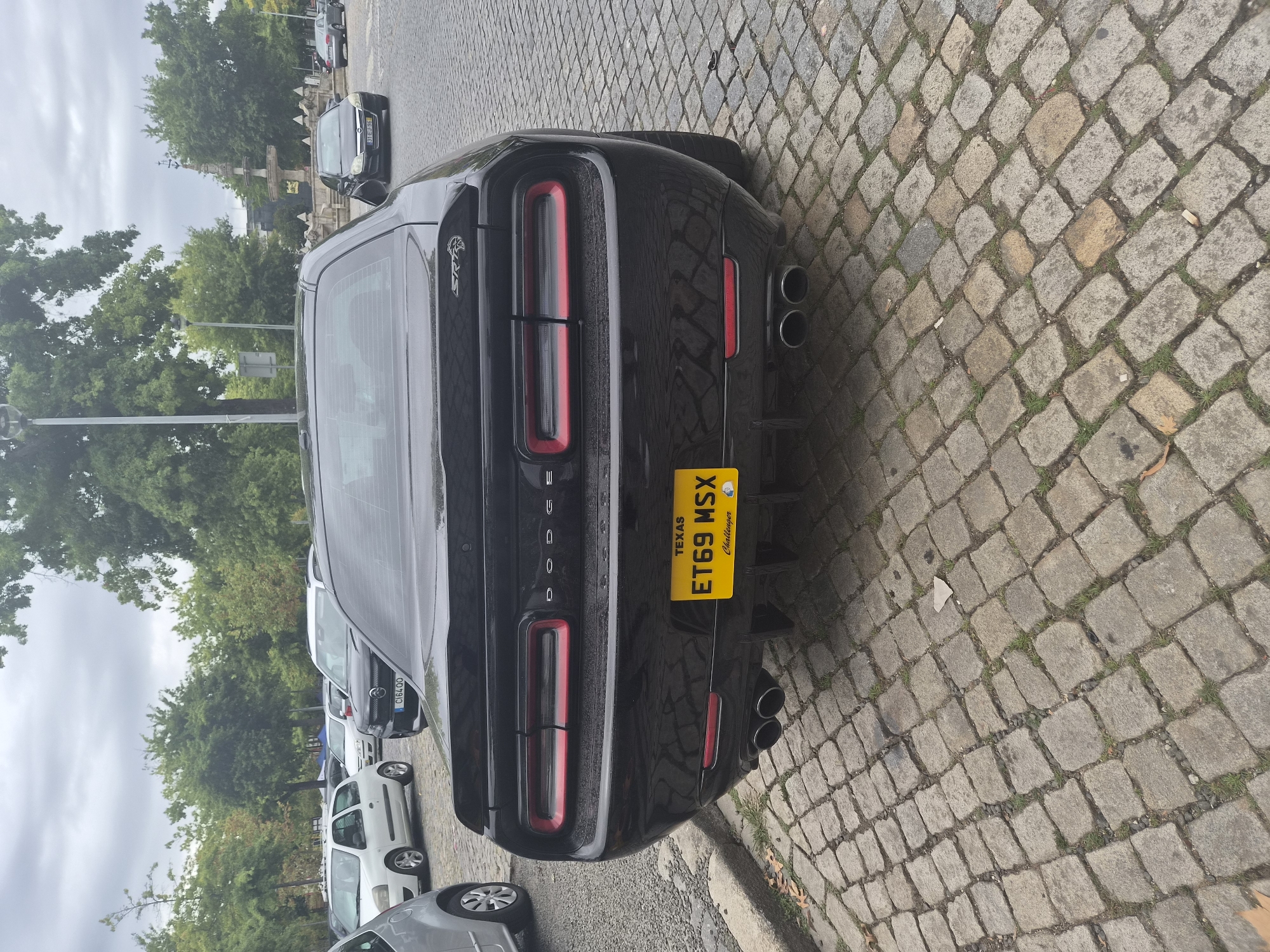 A black Dodge Challenger SRT is parked on a cobblestone street in Barcelos, Portugal. The car's rear end is prominent in the foreground, showcasing its taillights, exhaust pipes, and a Texas license plate reading "ET69 MSX". The car is positioned on a cobbled street with grass growing between the stones. In the background, trees and other parked cars are visible, suggesting a public parking area or a street. The overall scene appears to be an outdoor setting in a city environment.