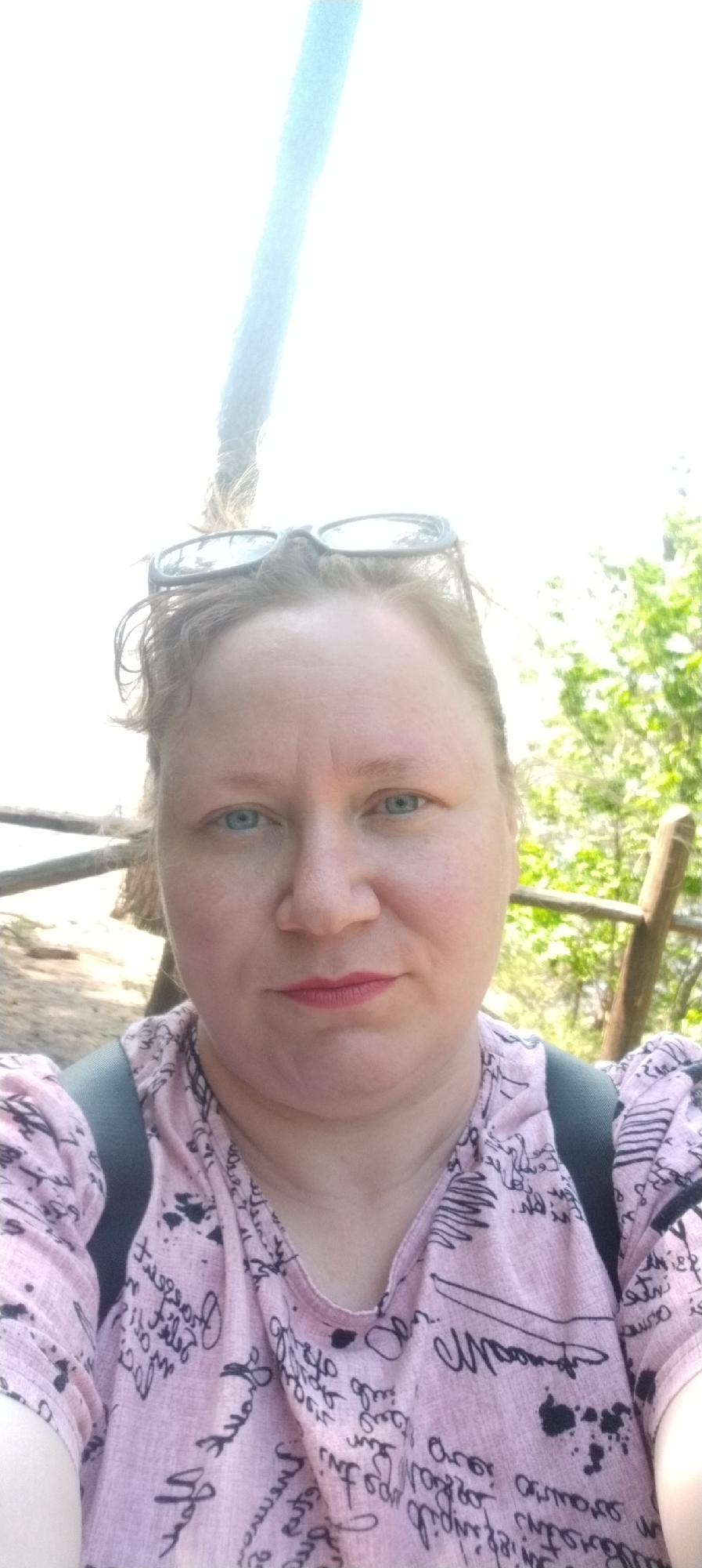 A selfie of a middle-aged woman with fair skin, blue eyes, and light-colored hair with sunglasses perched on top of her head. She is wearing a pink shirt with black writing. She appears to be outdoors, with a wooden fence visible in the background and trees and foliage to the right. The location is Noviy, Russia, though there's insufficient information in the image to confirm a precise address.