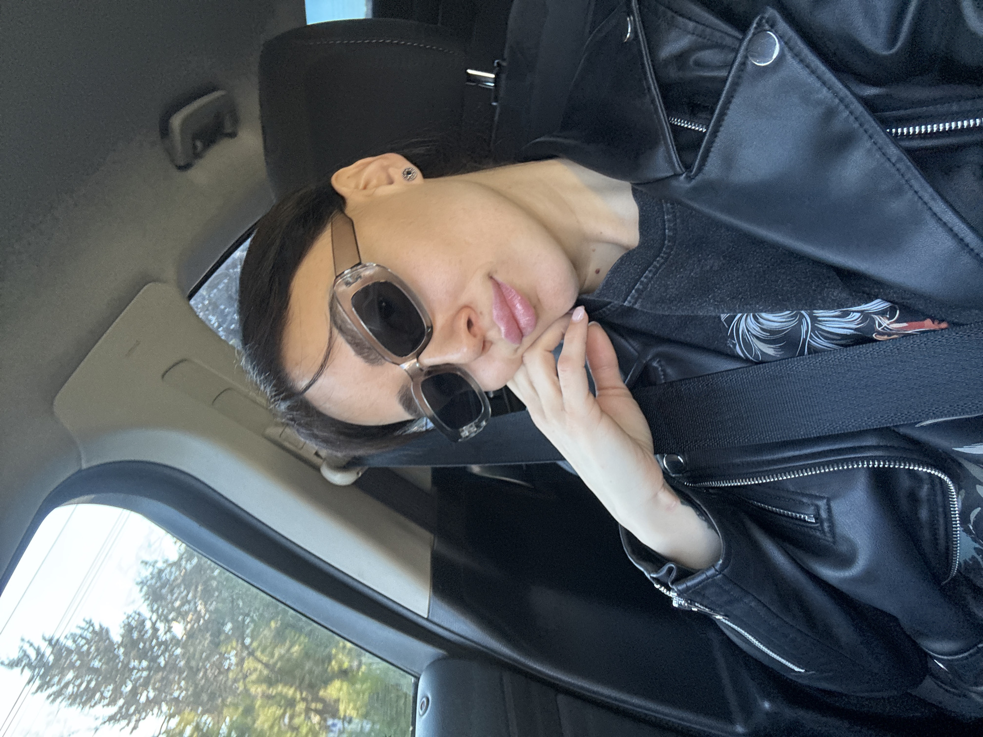 A woman is sitting in a car, wearing sunglasses and a black leather jacket. She is wearing a seatbelt and holding her hand to her face. Outside the window, trees can be seen. The location appears to be Tbilisi, Georgia based on the context of the user.