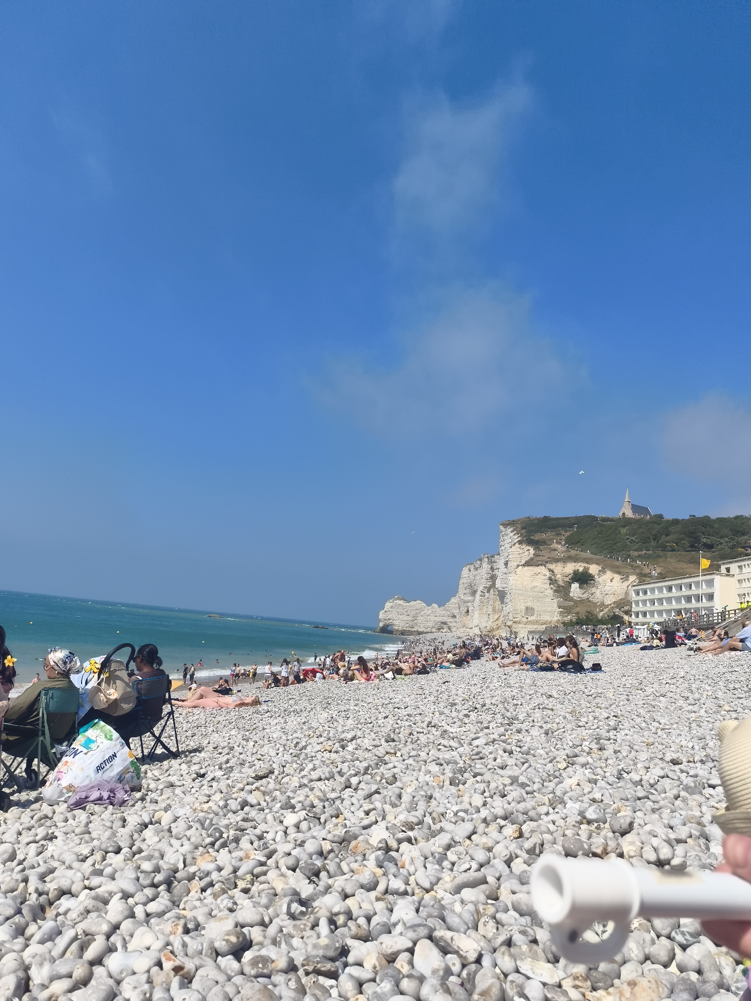 The image depicts a crowded pebble beach in Étretat, France, under a bright blue sky with sparse clouds. The sea is a vibrant turquoise, with small waves lapping at the shore. In the background, majestic white cliffs rise steeply from the coastline, with a church and buildings perched atop one of the cliffs. Numerous people are spread across the beach, some sunbathing on towels, others sitting in chairs, and many wading or swimming in the water. Beach umbrellas and bags are scattered amongst the pebbles. In the foreground, a person's hand holds a white cylindrical object. The overall scene is one of a bustling summer day at a popular coastal resort.