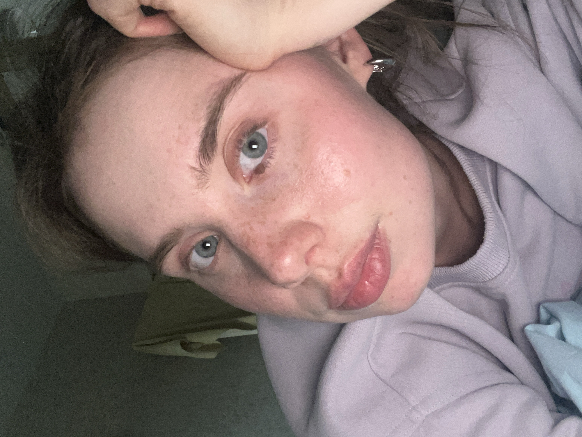 A close-up, indoor selfie shows a woman with fair skin, blue eyes, and freckles. She is wearing a light purple sweatshirt. Her head is tilted, with her hand near her forehead. There is a glimpse of a possible bed or surface with light-colored material in the lower portion of the frame. The exact location in the city of Chaikovsky, Russia cannot be determined from the image.