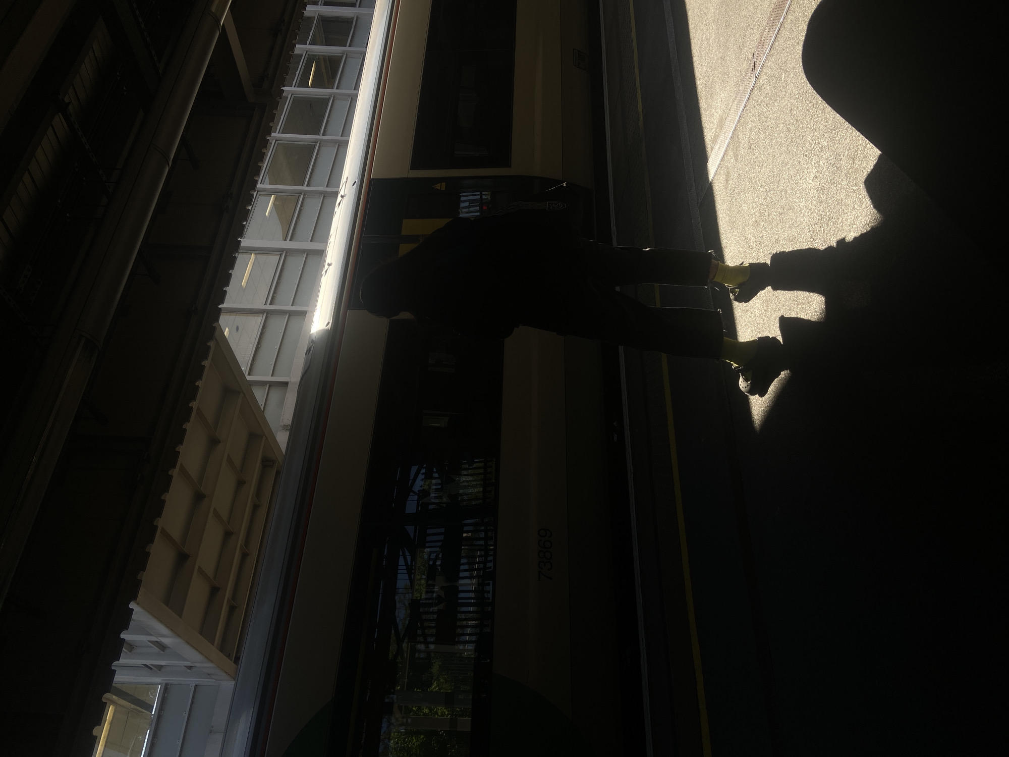 The image captures a high-contrast scene at a train station platform, bathed in strong, low-angle sunlight, suggesting either early morning or late afternoon. The perspective is slightly angled, looking towards a stationary train and a person on the platform.
The central subject is a person, rendered as a dark silhouette, standing beside a train carriage. The bright sunlight illuminates their lower legs and feet, revealing bright yellow socks or footwear. Their elongated shadow stretches dramatically across the sunlit platform.
To the left, a train is stopped at the platform. The visible side of the train is light-colored, possibly beige or white, with a darker lower section. Numerous rectangular windows line the upper part of the train carriage, some reflecting the bright sky or station infrastructure. The number "73869" is faintly visible on the side of the train.
The platform floor is a light, textured surface where the sunlight hits, contrasting sharply with the deep shadows covering most of the ground. Yellow safety lines are visible along the edge of the platform next to the train. Overhead, parts of the station structure are visible in shadow, including what appears to be a covered area or roof with grid-like patterns and some dark, structural beams or pipes.
The lighting creates a stark division between light and shadow, with much of the upper and right portions of the image being in deep shadow, emphasizing the solitary figure and the train caught in the sun's glare.