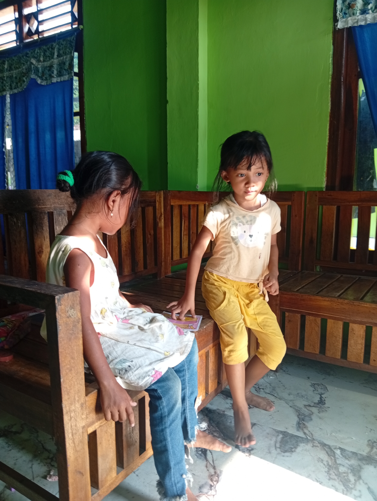 Two young girls are seated on a wooden bench in a room. The girl on the left is wearing a white sleeveless dress with a floral print and blue jeans. Her hair is tied up in a bun with a green accessory. The girl on the right is wearing a yellow t-shirt with a graphic print and yellow pants. Both girls are looking in different directions, and the girl on the right appears to be holding a small card or object. The room has green walls and wooden furniture, with sunlight streaming in from the left, casting shadows on the floor. The floor is tiled with a marble-like pattern. The scene appears to be indoors, possibly in a residential setting in Sri Minosari, Indonesia.