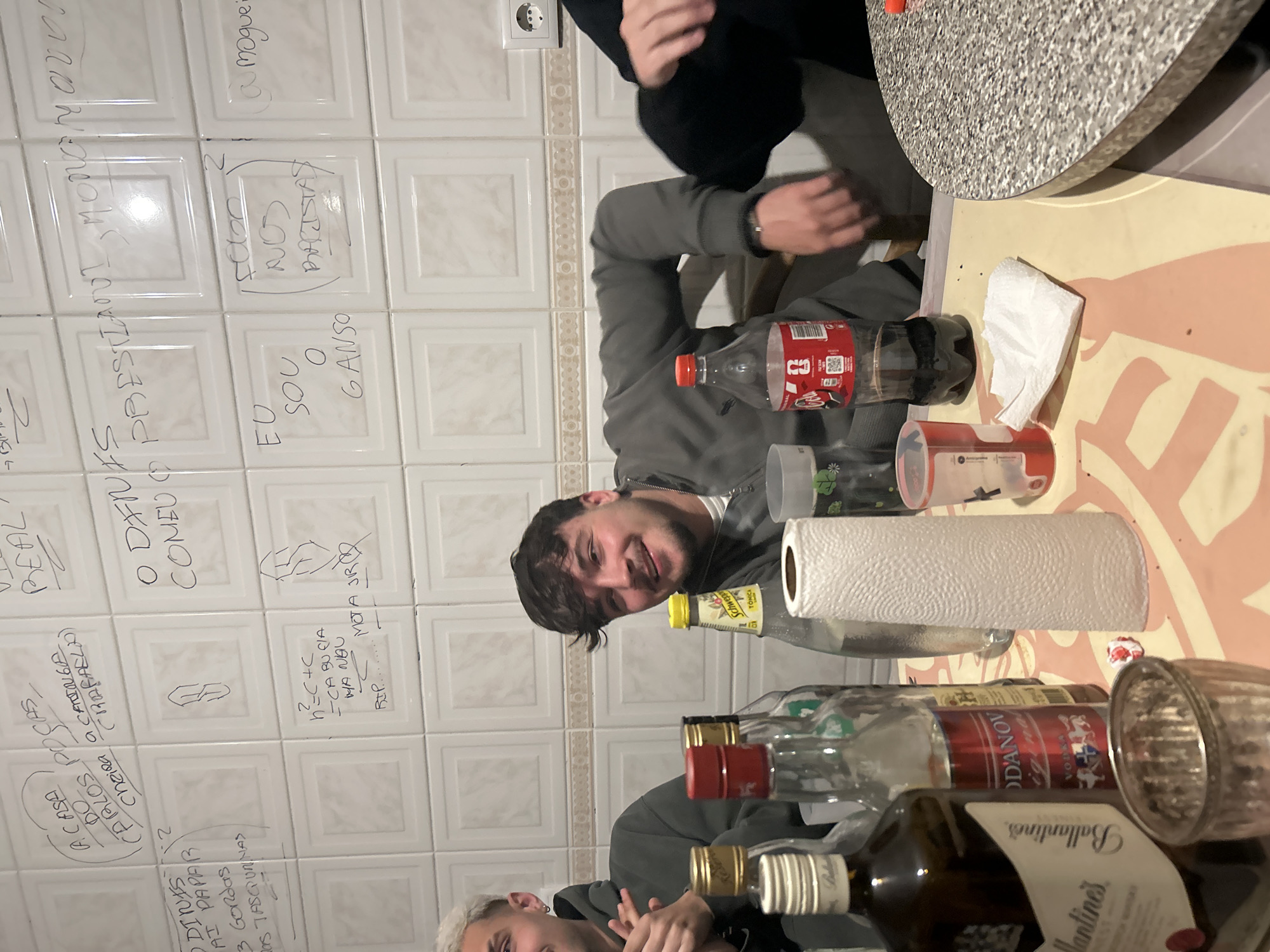 The scene depicts an indoor social gathering, identified as being in the city of Bragança, Portugal. Three individuals are partially visible around a light-colored table covered with a patterned surface.
In the center, a young male with dark, short, wavy hair and light facial stubble wears a grey zip-up hoodie over a white undershirt. He is smiling, leaning back in a chair, and looking towards the camera. To his left, another young male is partially visible; he has short, bleached blonde hair, an earring in his left ear, and wears a dark grey hoodie. His hands are raised and clasped together, suggesting clapping or a gesture. To the right, only the left arm and leg, clad in black clothing, of a third individual are visible.
The table in the foreground holds various items. A partially full dark brown bottle of Ballantine's Finest Scotch Whisky stands prominently. Beside it is a partially full clear bottle of Odanov Original Vodka with a red label. A clear bottle of Schweppes Tónica with a yellow cap and a clear plastic bottle of Coca-Cola are also present. A white roll of paper towels stands upright. Other objects include a crumpled white paper napkin, a silver-colored ridged glass, and a clear plastic cup with red markings, partially filled. The tabletop consists of a light-colored, possibly beige or pale pink, patterned surface, with a grey speckled section visible on the right.
The background consists of a tiled wall with square white tiles featuring a raised central square design. The grout lines are light brown. The tiles are extensively covered in handwritten graffiti in black marker, primarily in Portuguese. Visible phrases include "A CASA DO CARLOS POUCAS CHEIRA A CANINGA (RAFAELA)", "O DINIS VAI PAPAR 3 GORDAS DAS TASCUNINAS", "EU SOU O GANSO", and "FOGO NOS BATISTAS". Several stylized "S" symbols are also drawn. A white Type F power outlet is visible on the right side of the tiled wall. Artificial lighting illuminates the scene.