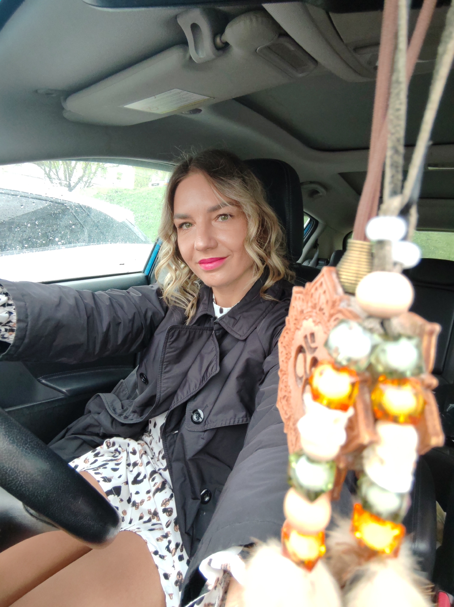 A woman is sitting in the driver's seat of a car. She is wearing a black trench coat over a white dress with black spots. She is holding the steering wheel with her left hand. There is a decorative hanging ornament in front of her. The car has a sunroof, and it appears to be raining outside. The location cannot be determined.