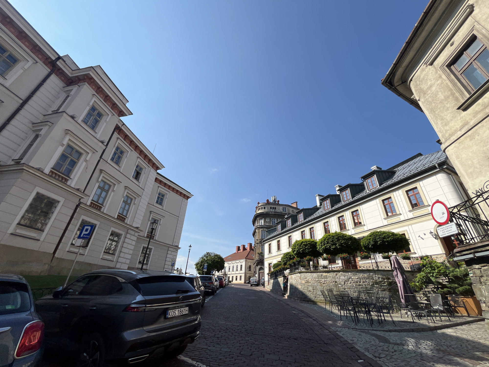 The media file depicts a sunlit street scene in a European town, likely during the day due to the clear blue sky and strong shadows. The perspective is low-angle, looking up at the architecture and along the cobblestone street.
The scene features several buildings with traditional European architecture. On the left, a large, light-colored building with multiple stories and red-trimmed windows dominates the foreground. To the right, a cream-colored building with a gray roof extends down the street, adorned with rows of windows and decorative trees planted along a stone wall. A distinctive circular tower structure is visible further down the street, integrated into one of the buildings.
Several cars are parked along the street, including a dark SUV in the foreground with a license plate that reads "KOS 1861H". There are also some outdoor tables and chairs set up, suggesting a cafe or restaurant. A sign with a red circle and a horizontal line, typical of a "no entry" or prohibition sign, is visible on the right side of the street, along with some Polish text.
The weather appears to be clear and sunny, creating sharp contrasts between light and shadow. The cobblestone street and the style of the buildings suggest an older, historic urban environment. No people are clearly visible in the image, and the overall mood is one of quiet daytime activity in a picturesque setting.