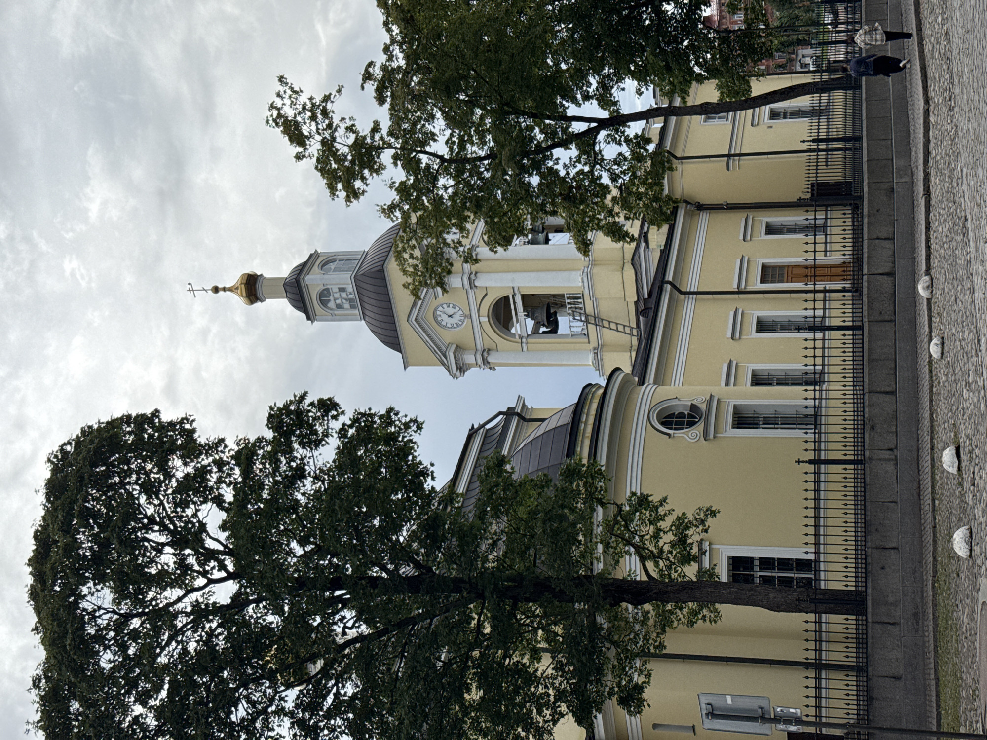 This image depicts the bell tower and dome of the Transfiguration Cathedral in Vyborg, Russia. The cathedral is painted a bright yellow and features white architectural trim. In the foreground, a black metal fence encloses the cathedral grounds. Beyond the fence, a cobblestone street is visible with two white bollards. Two individuals are walking away from the camera on a sidewalk to the right of the frame. Several large trees with dark green foliage are situated in front of and to the sides of the cathedral, partially obscuring the view of the building. The sky is overcast with grey clouds.