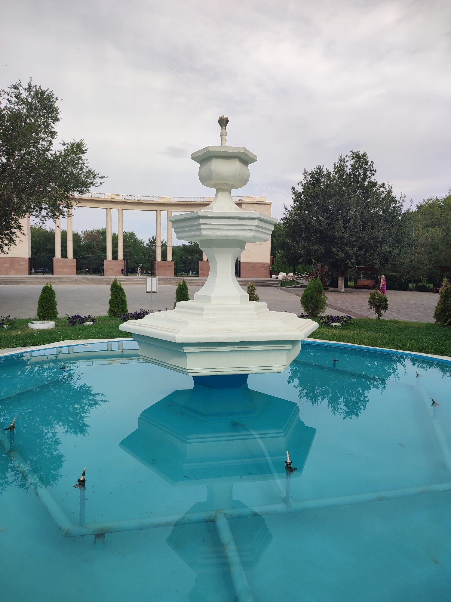 The image captures a white, multi-tiered fountain in the foreground, its lower octagonal section reflected in the vibrant blue water of the basin. The basin itself appears somewhat aged, with visible pipes and nozzles beneath the water's surface. Beyond the fountain, a paved area leads to a colonnade with evenly spaced, tall, light-colored pillars. In the background, lush green trees and manicured lawns suggest a park-like setting. A few people are visible in the middle ground, one walking near the colonnade and another in the distance near a wooden bench. The sky is overcast with a light gray hue. The overall scene is set in the city of Qonaev, Kazakhstan, likely within a public park or square designed with classical architectural elements.