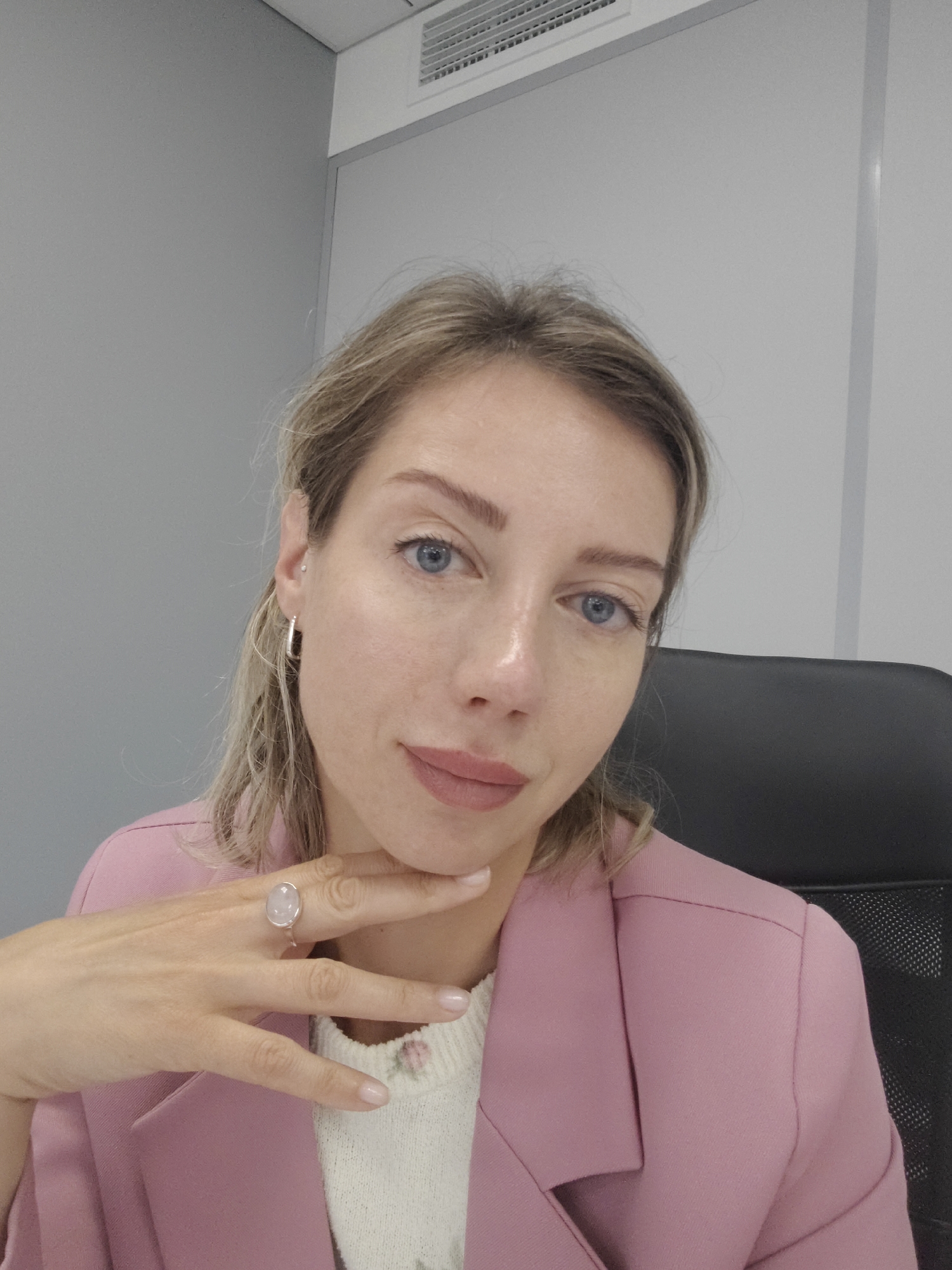 A blonde woman with blue eyes and fair skin is pictured sitting in an office chair. She is wearing a pink blazer over a white sweater with a floral pattern. Her left hand is resting on her chin, and she is wearing a ring with a large pink stone. The background is a plain gray wall and a white ceiling with an air vent. The image appears to be a selfie, taken from a slightly high angle. The woman is looking directly at the camera. The lighting is even and appears to be natural. There are no other people or objects in the frame that provide specific information about the location within Saint Petersburg.