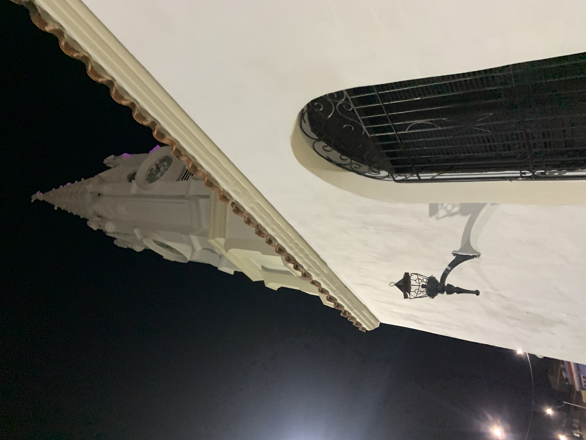 An upward-tilted view at night captures architectural details in Juan Griego, Venezuela. A white building with a tiled roof overhangs the dark sky. A window with a decorative black wrought iron grate is visible on the side of the building. A black lantern extends from the side of the building. In the background, a partially visible church with a tall clock tower is also seen. Streetlights are present at the bottom of the frame.