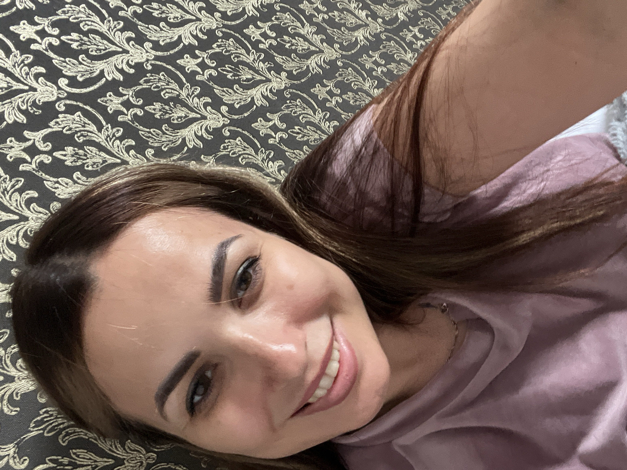 The media file is a close-up, overhead shot of a young woman lying on her side on a patterned surface. She is looking towards the camera and smiling. Her hair is long and brown, falling around her face and shoulders. She is wearing a light purple garment, possibly a hoodie or a sweater, which is draped around her.
The surface she is lying on is dark and features a repeating gold damask-like pattern. This surface appears to be a bedspread or a couch. The lighting suggests it is daytime, possibly indoors, as there are no strong shadows. The woman's expression is happy and relaxed, indicated by her smile and the crinkling around her eyes. Her makeup includes defined eyebrows and eyeliner. The image is sharp and clear, with a shallow depth of field that blurs the background slightly. There is no visible text in the image.