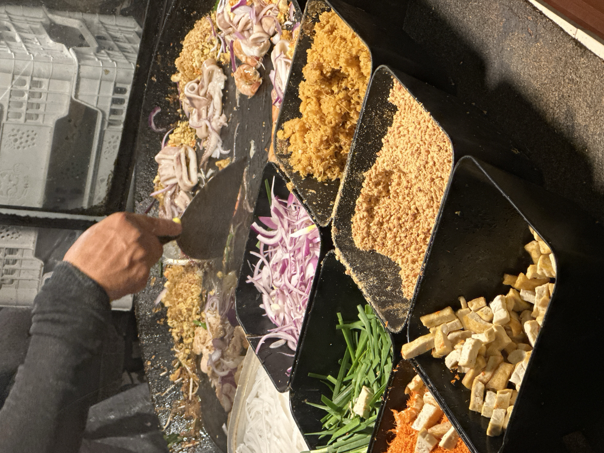 A bird's-eye-view shot shows a person cooking a street food dish in Pattaya City, Thailand. The cook, seen with a bare hand and part of an arm clad in a dark sweater, is using a flat spatula to mix ingredients on a flat cooking surface. Scattered around the cooking area are several black containers holding various ingredients. These include cooked squid, fried yellow bits, ground peanuts, thinly sliced red onions, green chives, and cubes of fried tofu. In the bottom corner, there is also a small container with orange shredded ingredients. The dish being prepared includes some clear, thin noodles. Some of these ingredients are mixed together on the cooking surface, while others are still separate. The background is dark and slightly out of focus, with a white, textured surface visible near the cooking surface. The lighting appears to be from overhead, casting shadows on the containers and cooking surface.