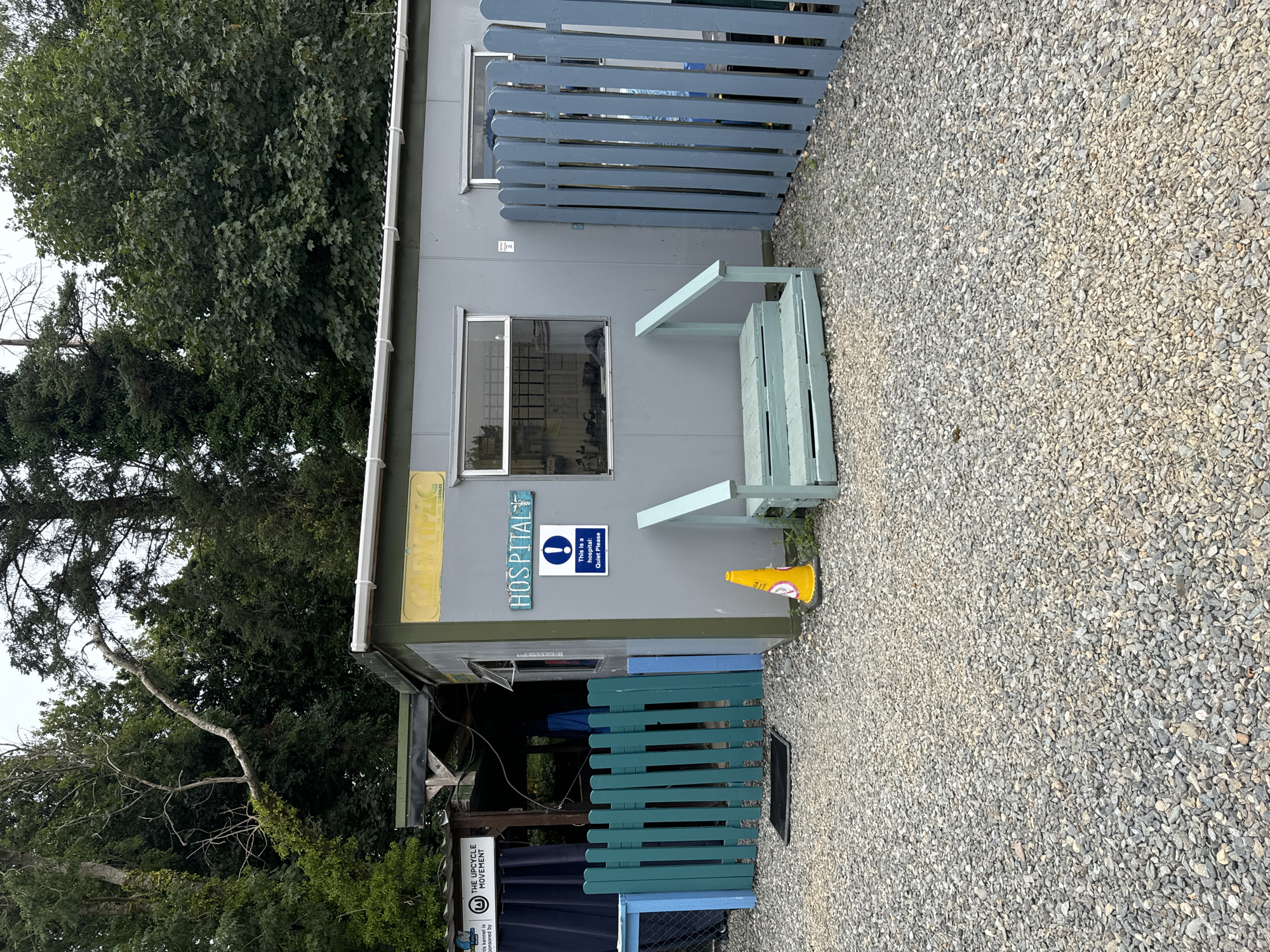 An outdoor scene in Courtown, Ireland, features a grey, portable building with a pitched roof, positioned on a gravel ground. The building has two windows with metal bars on the left, and a wider window on the right. A blue sign above the right window reads "HOSPITAL" in white lettering with a caduceus symbol. Below this sign, a blue and white sign displays an exclamation mark symbol and the text "This is a hospital: Quiet Please". A short, light blue wooden staircase leads up to the building's entrance. A yellow traffic cone is placed on the gravel to the right of the entrance. To the left of the building, a turquoise wooden fence with blue posts encloses an area with a black mat on the ground. In the background, dense green trees and foliage are visible under an overcast sky. A portion of a sign for "THE UPCYCLE MOVEMENT" is visible on the left.
