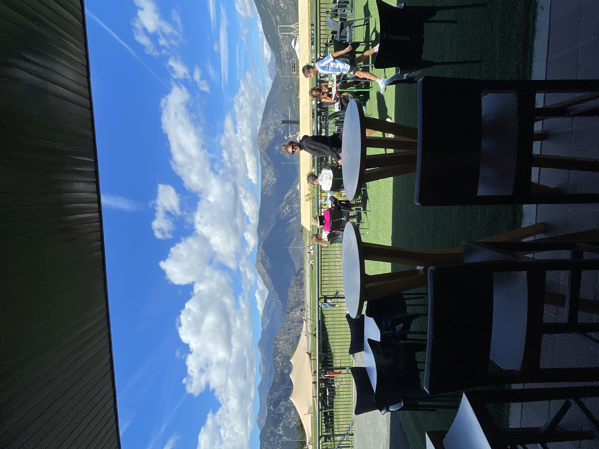 The image depicts an outdoor seating area, likely a patio or terrace, with tables and chairs set up on a green, turf-like surface. In the background, a large, beige, tent-like structure is visible, possibly for an event or market. Beyond that, a range of mountains forms the horizon, with a bright blue sky dotted with white clouds above. Several people are seated at tables or walking in the mid-ground. In the foreground, two round tables with wooden legs and grey tabletops are prominent, along with dark chairs. A woman wearing sunglasses and a black hoodie is standing near one of the tables, looking towards the camera. A young boy in a striped shirt is walking past another table. The scene suggests a relaxed outdoor atmosphere, potentially a cafe or restaurant in a scenic location. Given the prominent mountain range and the style of the tent, this scene could be located in Juberri, Andorra, a region known for its mountainous landscapes and outdoor activities.