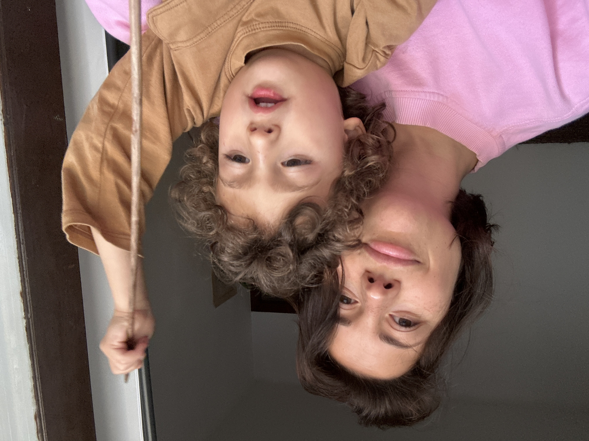 The image shows a woman and a child posing upside down. The child is on top, wearing a brown shirt. The woman is below, wearing a pink sweatshirt. They are likely inside a building, near a window or doorway. A dark-colored frame is visible. The image does not provide any information to verify the location is Barcelona, Spain.