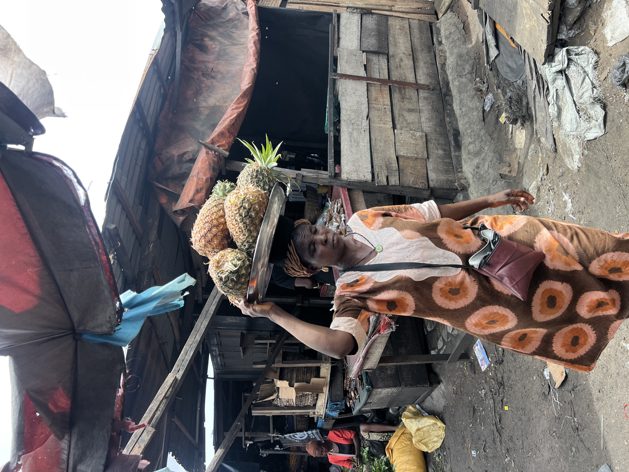 In Nkpor, Nigeria, a woman carries a silver tray piled with pineapples on her head. She wears a patterned brown and white dress and a small, dark-red purse on her shoulder. The environment includes makeshift buildings constructed of wood and tarps. Debris litters the ground. Another person sits on the ground nearby.