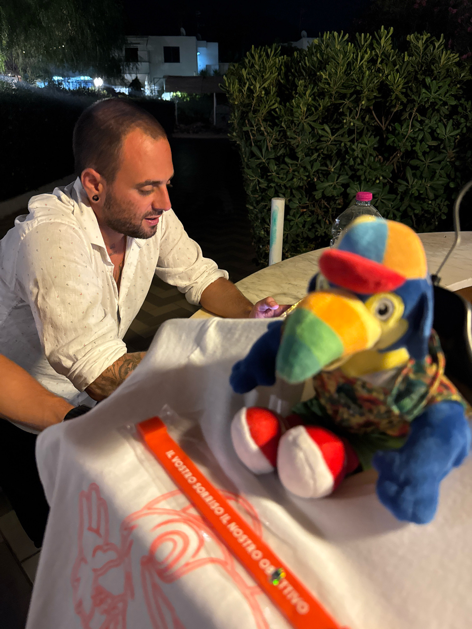 In a dimly lit outdoor setting at night, a man with short hair, a beard, and an earring is seated at a table. He is wearing a white, patterned, open-collared shirt. In front of him, a stuffed parrot toy with a colorful hat and scarf sits on a white cloth. An orange band with Italian text "IL VOSTRO SORRISO IL NOSTRO..." is visible on the cloth. Behind the table, a hedge of green bushes is illuminated, and a building with lights is visible in the background. The scene is consistent with an outdoor dining area in a town like Isola delle Femmine, Italy, where natural vegetation and nearby buildings are common features.