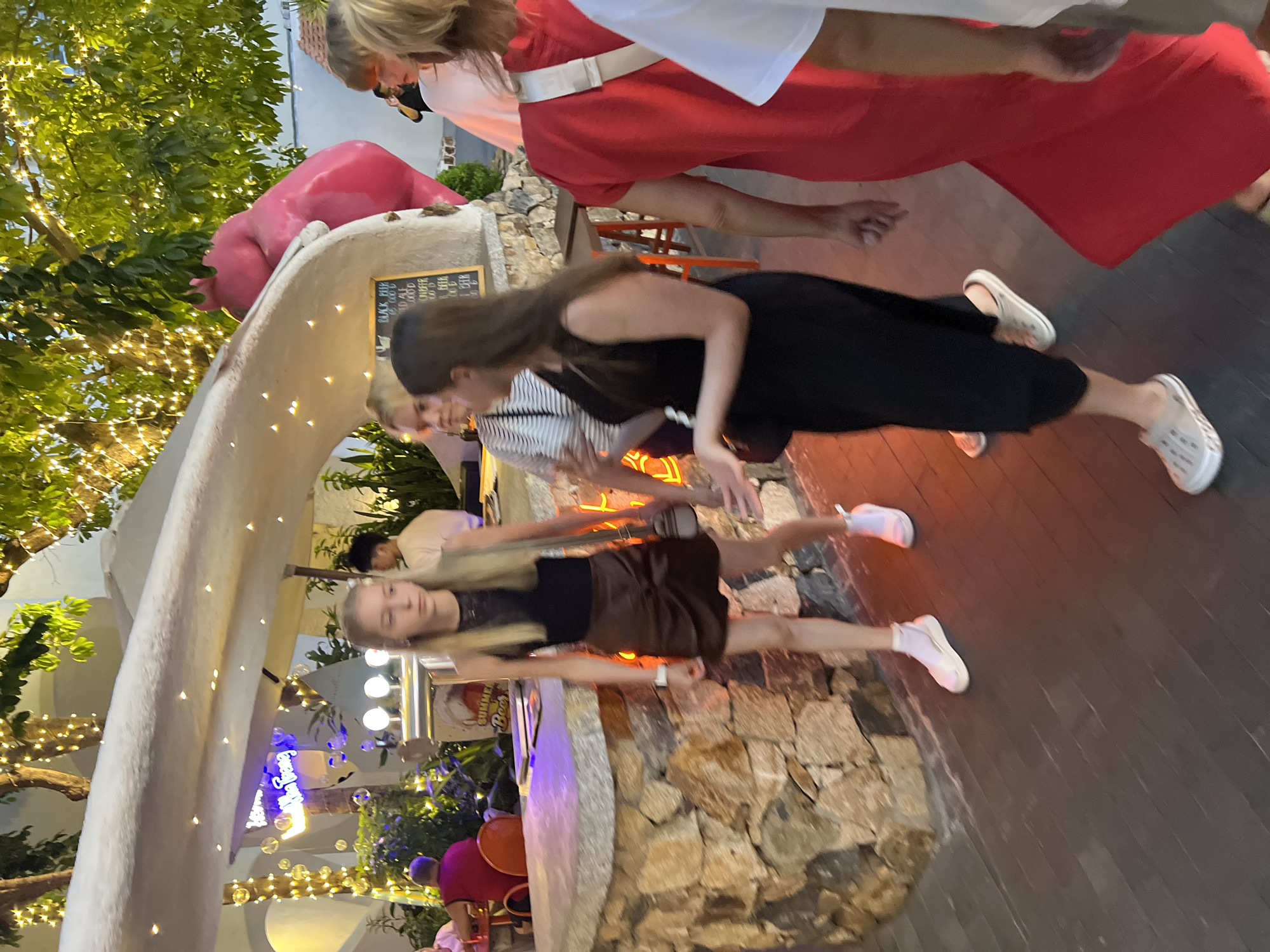 A group of people are gathered outdoors near a building with bright decorative lighting. In the foreground, a blonde woman in a black top and brown skirt walks towards the right, holding a dark purse. To her left, a woman with dark hair in a black dress walks in the same direction, also holding a dark purse. Behind them, another woman in a striped top is visible. Further back, a man in a white shirt stands near a bar area. A prominent pink sculpture of a hippopotamus is positioned on a curved white structure in the background. The environment appears to be a lively outdoor venue, possibly a bar or restaurant, with stone and wooden elements visible. The lighting suggests it is evening. The image is set in Nha Trang, Vietnam, as indicated by a neon sign that partially reads "Nha Trang".