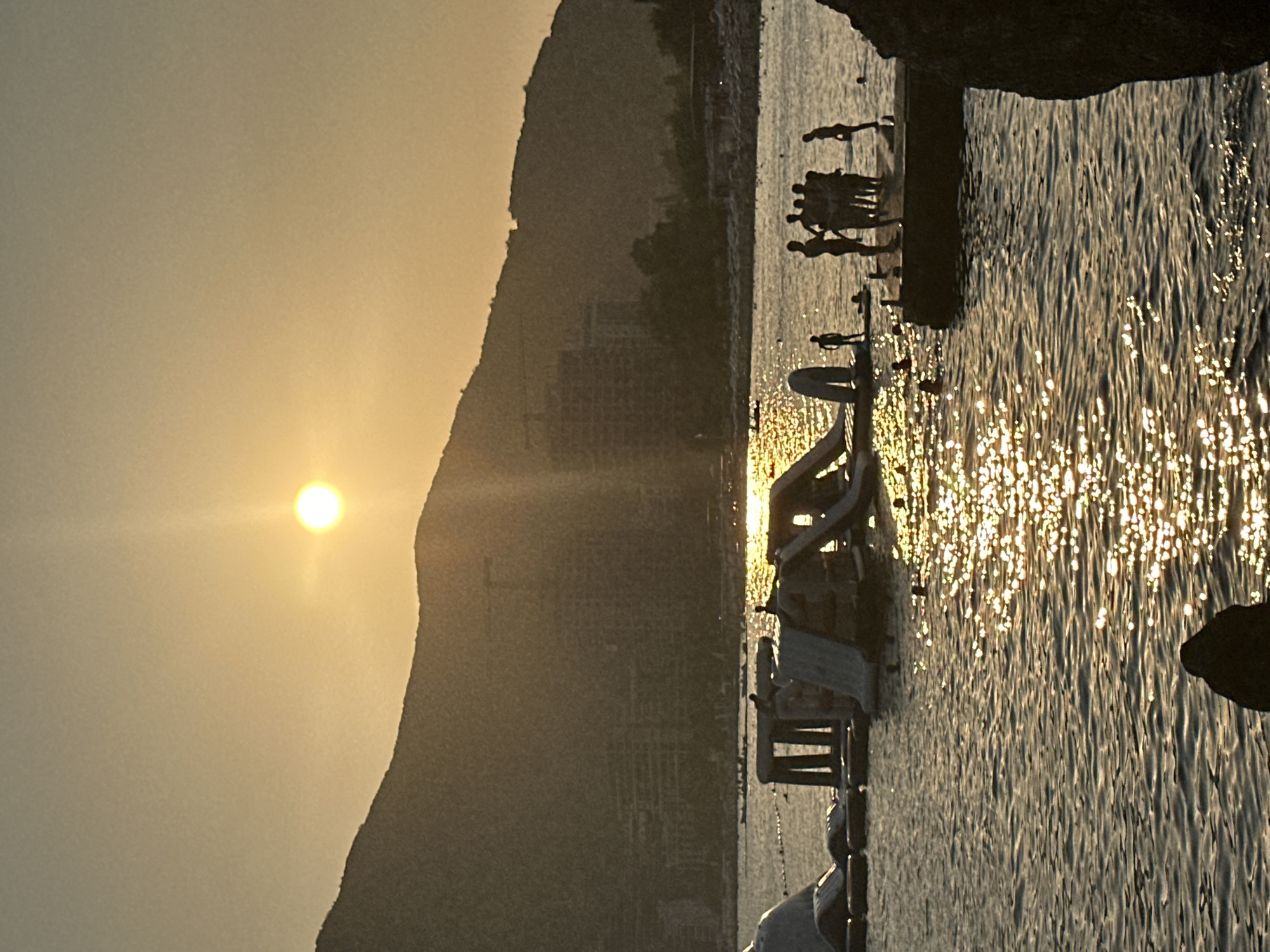 The image depicts a sunset scene at a beach in Budva, Montenegro. In the foreground, the Adriatic Sea is visible, with the setting sun casting a golden shimmer on its surface. Silhouettes of people are present, some standing on a pier and others in the water. To the left, an inflatable water park with slides and structures is partially submerged in the water. In the background, a mountainous landscape forms the backdrop, with buildings and construction cranes indicating a developed urban area. The overall atmosphere is serene and captures a summer evening at the coast.