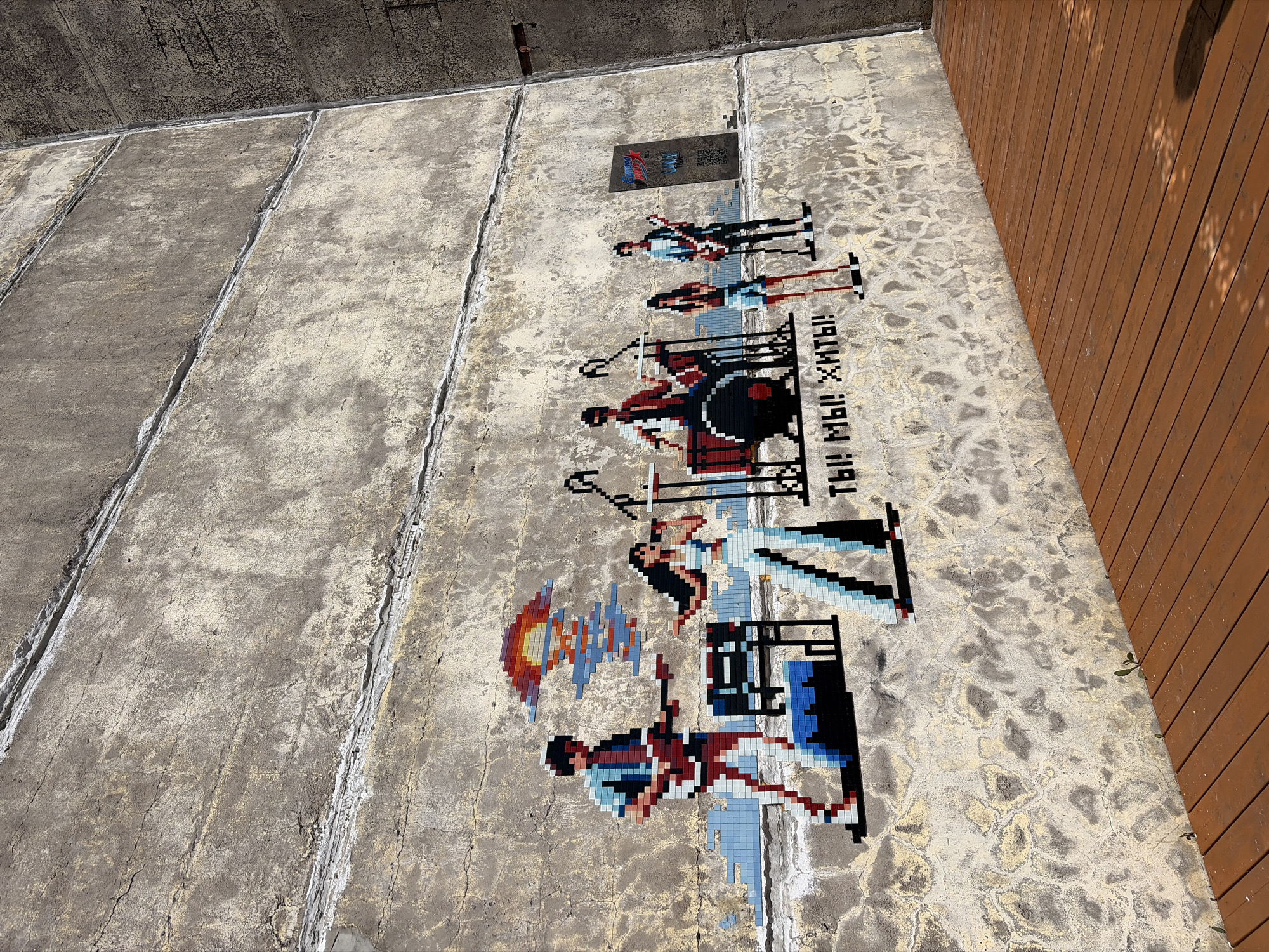 A large concrete wall, textured and aged with numerous cracks and horizontal seams, features a colorful pixel art mural. The mural depicts a four-member band performing. From left to right, the band consists of a male guitarist in a blue and red striped shirt, a female vocalist in a light blue top and white pants standing next to a keyboard stand, a male drummer behind a red and black drum kit, and a second male guitarist in a blue and black striped shirt. Above the central band members, a pixelated motif resembles either a vibrant sunset or an abstract explosion of red, orange, and blue. Below the central figures, the Cyrillic text "ТЫ! МЫ! ХИТЫ!" (You! We! Hits!) is rendered in black and white pixels. To the far right of the mural, a rectangular black poster with the "Europa Plus" logo and a QR code is affixed to the concrete wall. The base of the wall meets a reddish-brown wooden boardwalk composed of parallel planks, extending into the foreground and towards the right. Small green plant sprouts emerge from cracks along the bottom of the concrete wall and between the boardwalk planks. On the far right, a darker, coarse concrete structure forms an interior corner with the main wall. The scene is brightly lit by natural daylight, casting some distinct shadows onto the wooden boardwalk. This artwork is located at Sevkabel Port in Saint Petersburg, Russia.