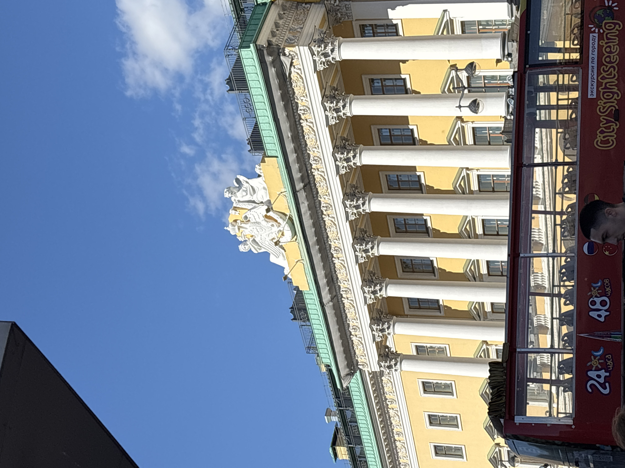 This image captures a bright, sunny day in Saint Petersburg, Russia, with clear blue skies and a few wispy clouds. The scene is dominated by the ornate yellow facade of a grand historical building, characterized by its numerous white columns and intricate decorative friezes. At the top of the building, a white sculptural group is visible.
To the right of the building, a bright red double-decker tour bus is parked, suggesting a tourist activity. The bus features prominent text on its side, including "City Sightseeing" in English, and "экскурсии по городу" in Russian, which translates to "city excursions." Other visible text includes numbers like "24" and "48" possibly indicating tour durations or prices, and the word "часов" (hours). There are also small flag icons, one appearing to be the Russian flag.
The angle of the photograph is unusual, tilted upwards and to the side, giving a dynamic perspective of the architecture and the tour bus. The sunlight casts distinct shadows, indicating it's likely daytime, perhaps late morning or early afternoon. The presence of people is hinted at with the upper body of a person visible in the bottom right corner of the bus's window, and another person partially visible at the very bottom right edge of the frame. The overall mood is one of vibrant urban exploration and sightseeing.