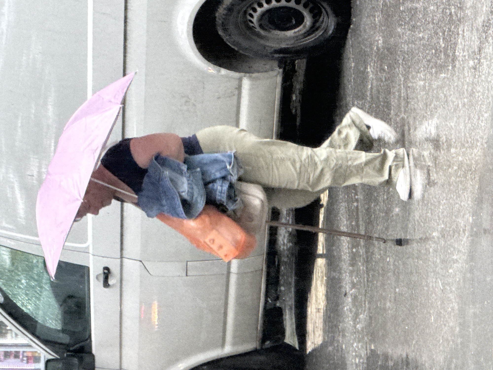 A person standing next to a white van in the city of Plovdiv, Bulgaria, is holding a pink umbrella and a walking stick. The person is wearing beige pants, a dark top, and white shoes. A folded blue jacket and an orange bag are draped over the person's arm. The van's front driver's side tire is visible, as is the vehicle door. The ground is wet and grey.