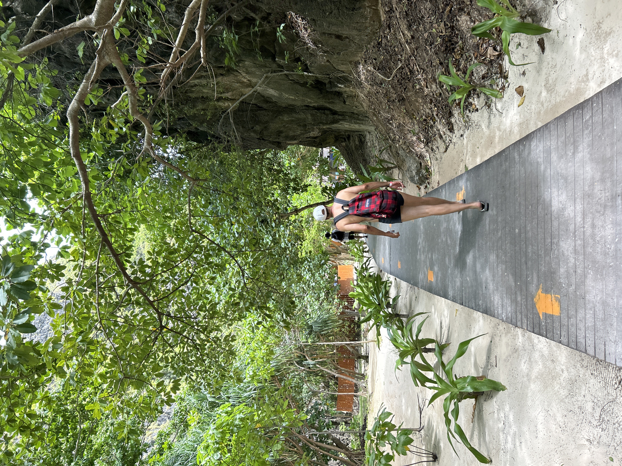 In Ao Nang, Thailand, a person walks along a wooden boardwalk lined with sand and lush green vegetation. The boardwalk has orange arrows painted on it, indicating a directional path. The person, seen from behind, wears a white cap, a red and black plaid backpack, and dark shorts. They are walking away from the camera towards a rocky cliff face on the right side of the frame. The environment suggests a tropical location with dense foliage and the characteristic limestone karsts of the region. Another person is visible further down the boardwalk in the distance.