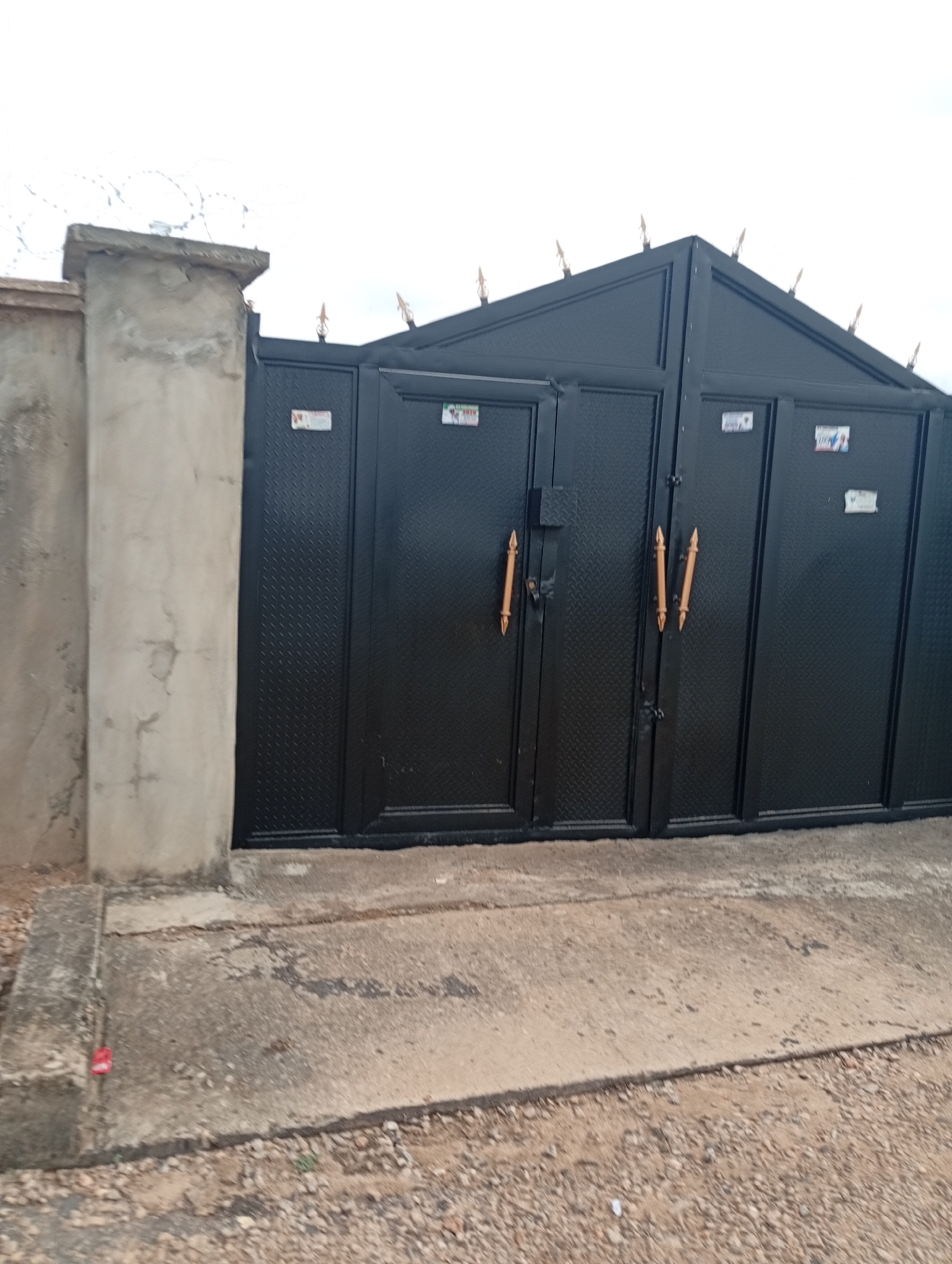 The image depicts a black metal gate in Adankolo, Nigeria. The gate is flanked by a concrete pillar to the left. The gate's design includes diamond-patterned panels and decorative spear-like points along the top edge. Two doors with golden, spear-shaped handles comprise the gate's central portion. Small stickers are visible on various gate panels. The ground is a mixture of concrete and gravel. Barbed wire can be seen atop the wall in the background.