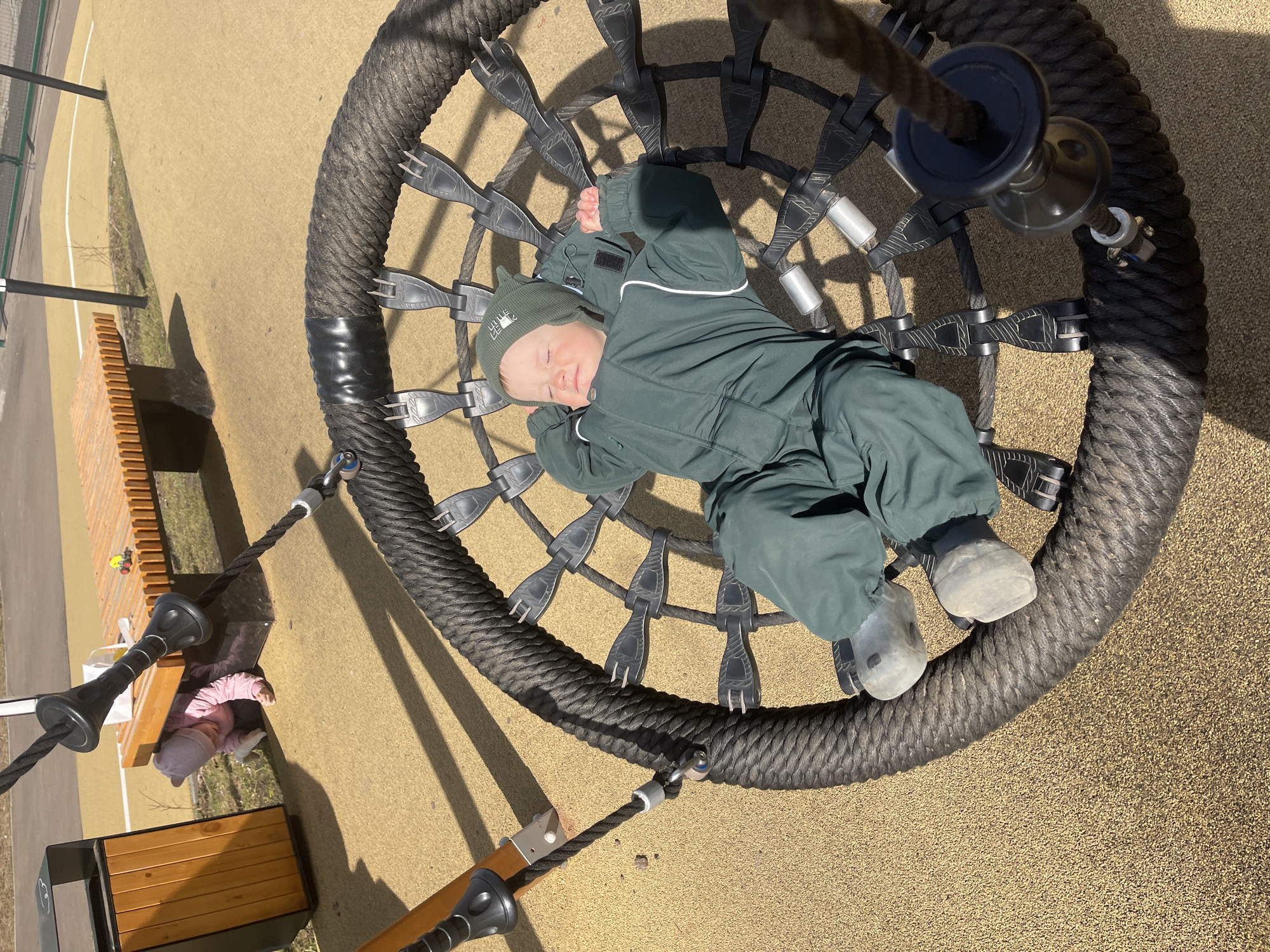 A young child sleeps on a large, circular rope swing. The child is wearing a green winter suit and a matching hat. The swing is located in a park or playground, possibly in Izhevsk, Russia, as indicated by the prompt. The swing's design features a thick, woven rope forming the outer ring, with a network of smaller ropes connecting to a central point. A bench and trash bin are visible in the background. The ground is covered with a tan rubber or composite safety surface.