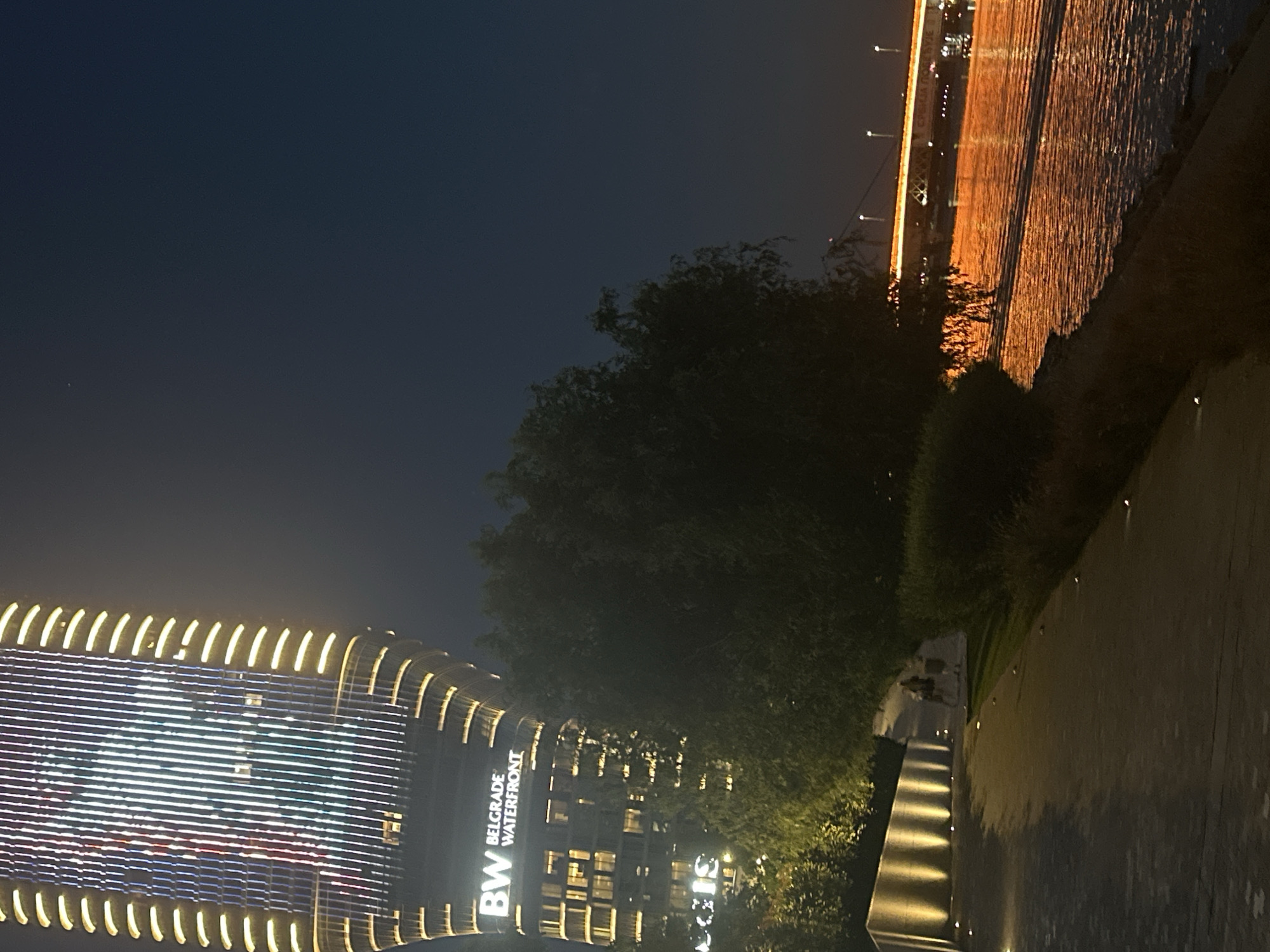 The image depicts a nighttime scene along the Belgrade Waterfront in Belgrade, Serbia. A tall, modern skyscraper with illuminated vertical lines of white, red, and blue lights dominates the left side of the frame. The text "BW BELGRADE WATERFRONT" is visible on the lower portion of the building.
In the foreground, a paved walkway curves along the riverbank, lined with low, embedded lights. Several people are walking on the walkway in the middle ground, appearing as silhouettes against the ambient light. To the right, a body of water, likely the Sava River, reflects the orange and yellow lights from a distant bridge, which spans across the water and is also illuminated. Trees and other vegetation are present along the riverbank, creating a natural element contrasting with the urban architecture. The sky is a dark, deep blue, indicative of night.
